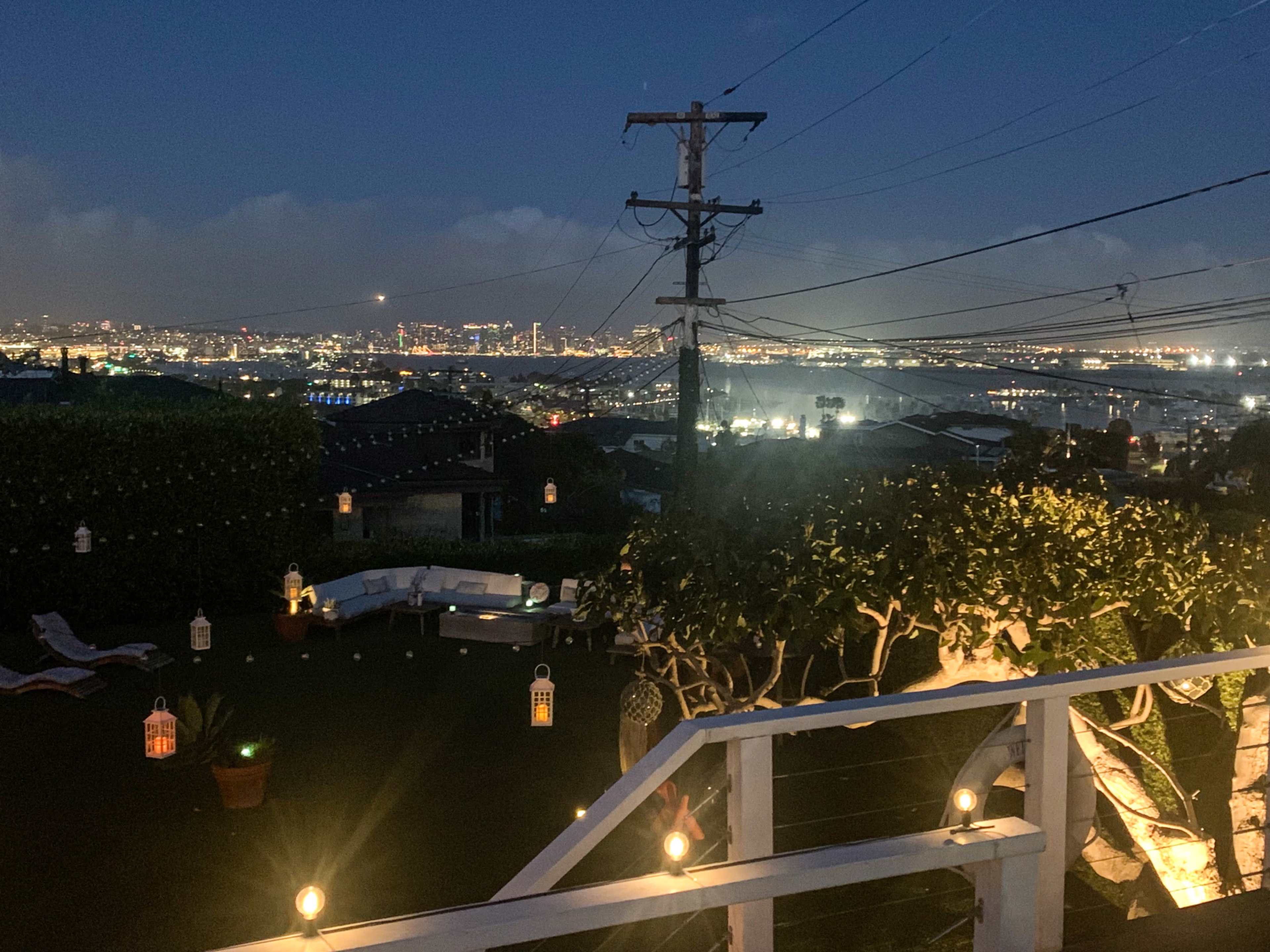 A view from a balcony overlooking a city skyline illuminated at night, with string lights and lanterns in a landscaped garden below.