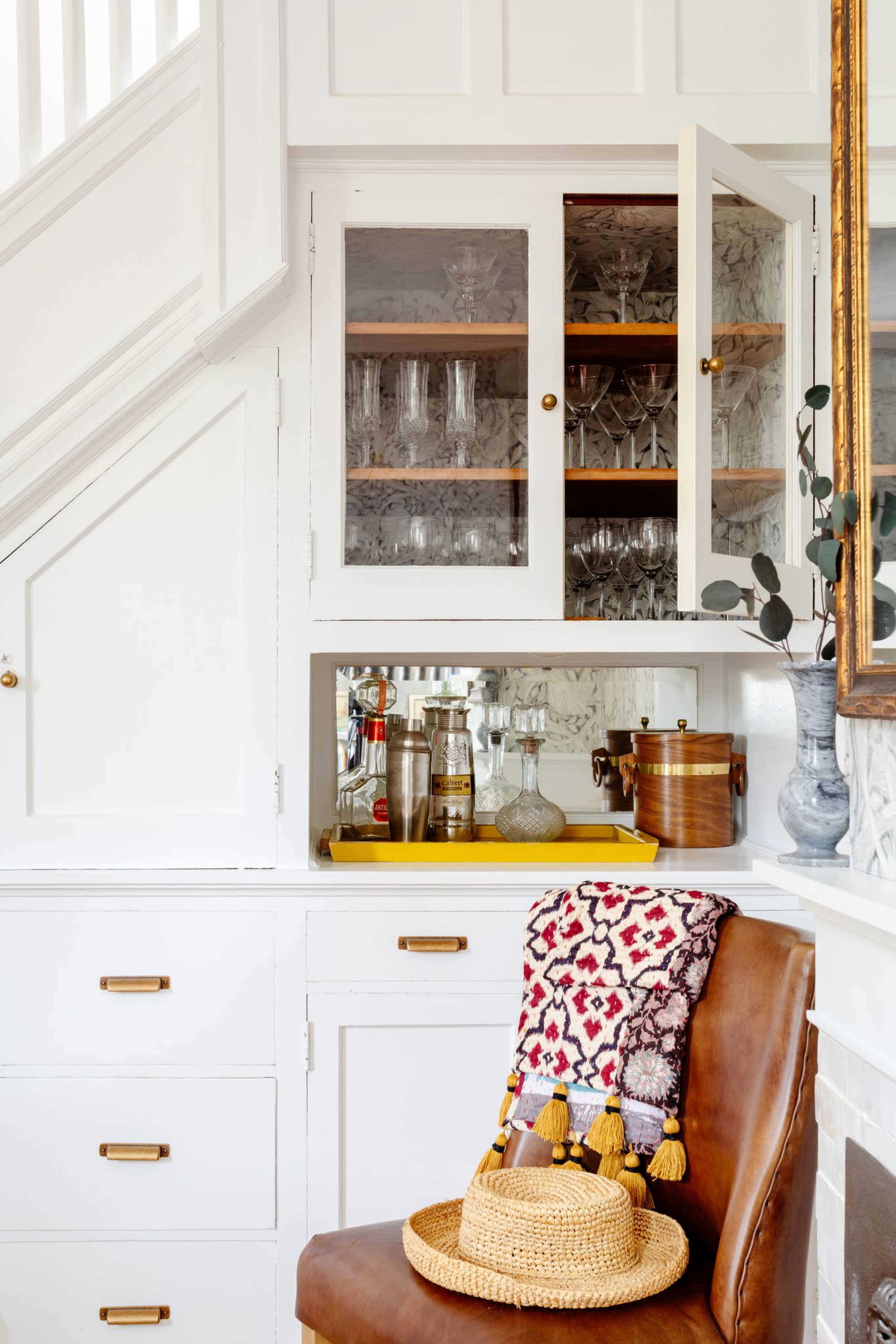 A corner of a kitchen features a white cabinet displaying glassware, a wooden bar cart with bottles, and a decorative chair draped with a colorful patterned blanket and a straw hat.
