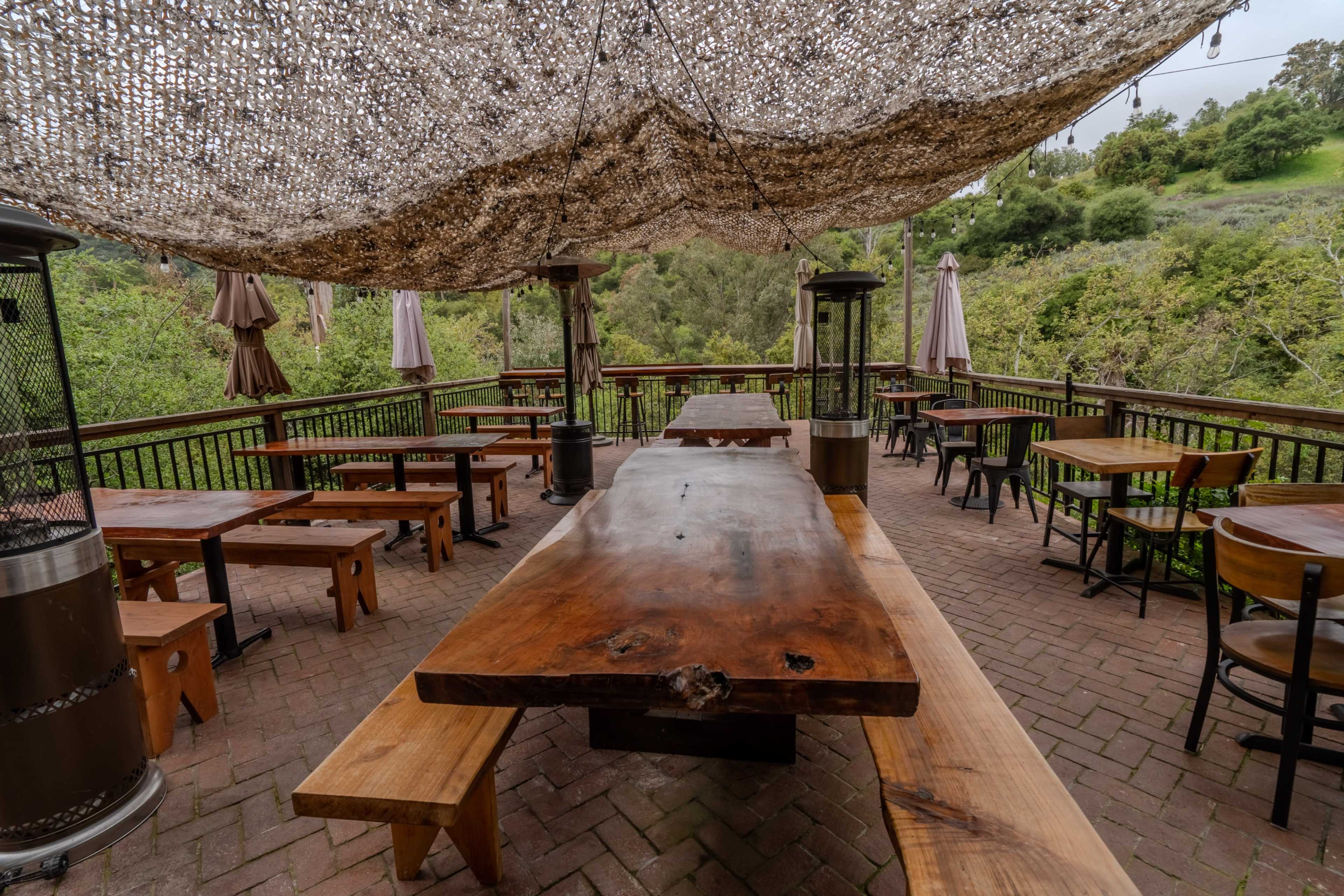 The image shows a wooden outdoor dining area with a large table, benches, and shaded by a canopy, surrounded by greenery.
