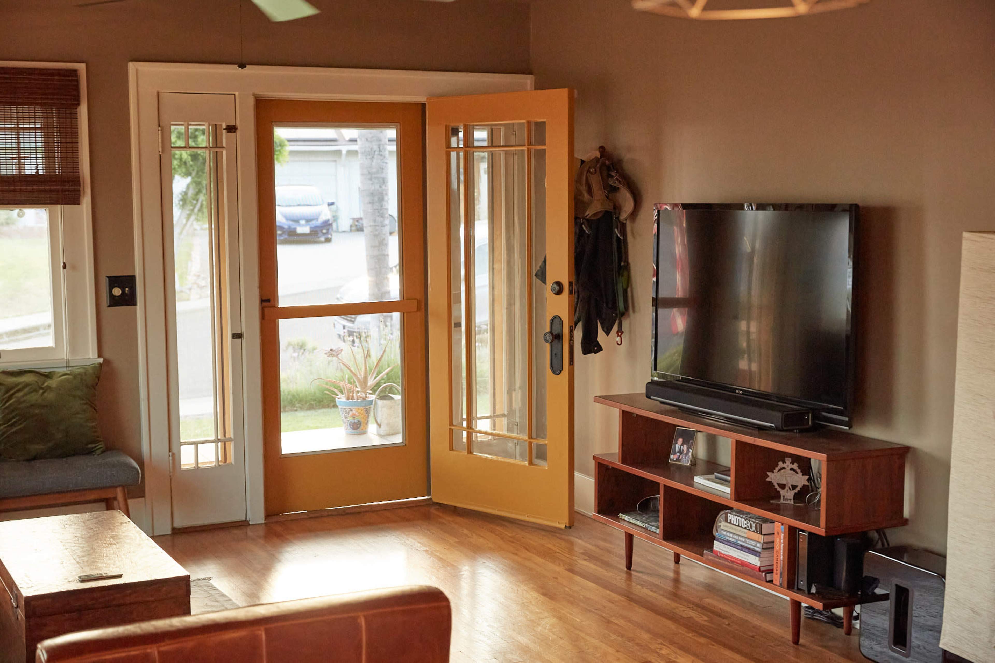 A living room with a yellow front door opening to the outside, a brown couch, a wooden coffee table, and a television on a wooden stand.