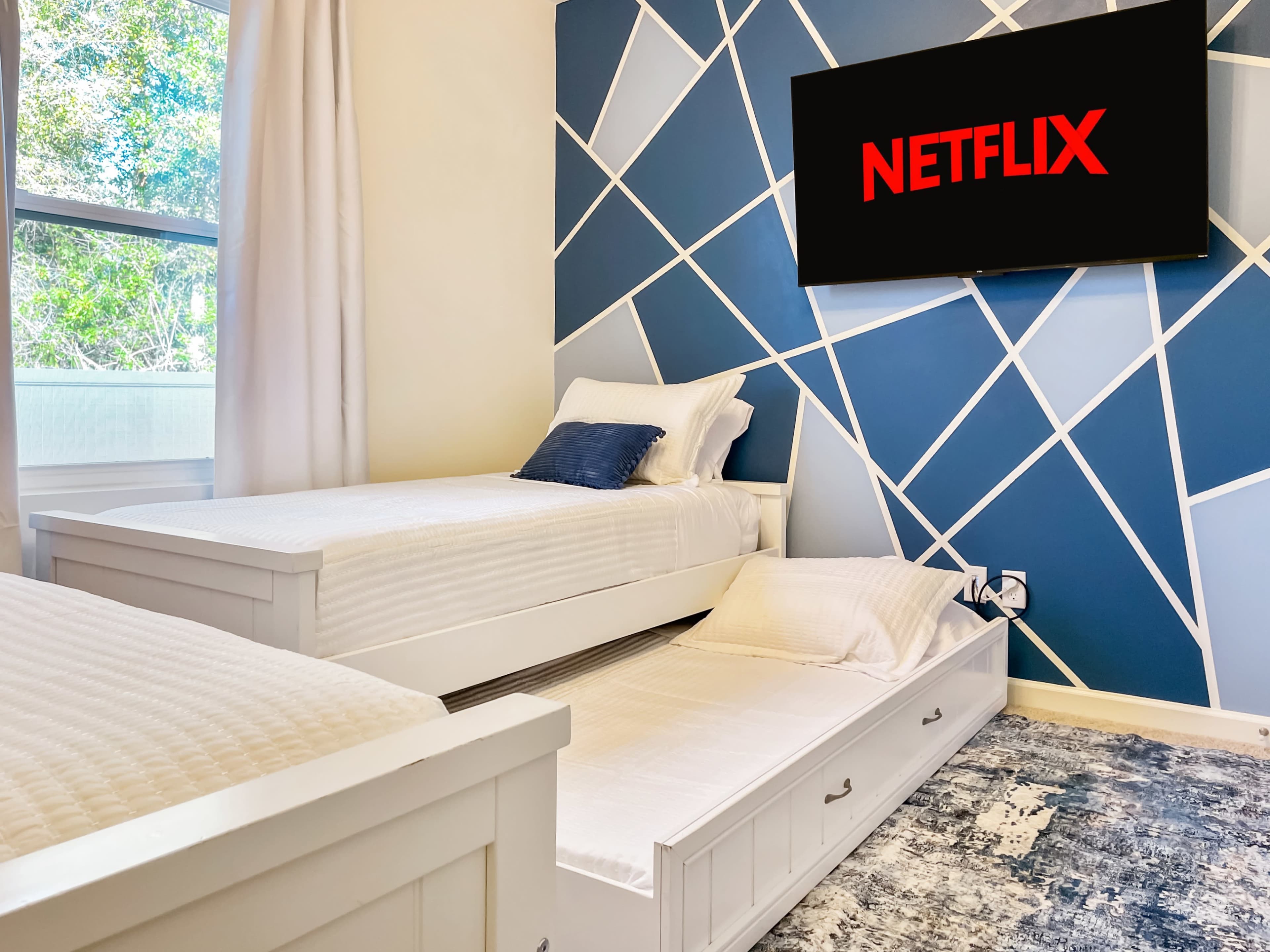 A bright bedroom features two white beds, one of which is a pull-out, with a large TV displaying the Netflix logo on a geometric blue accent wall.