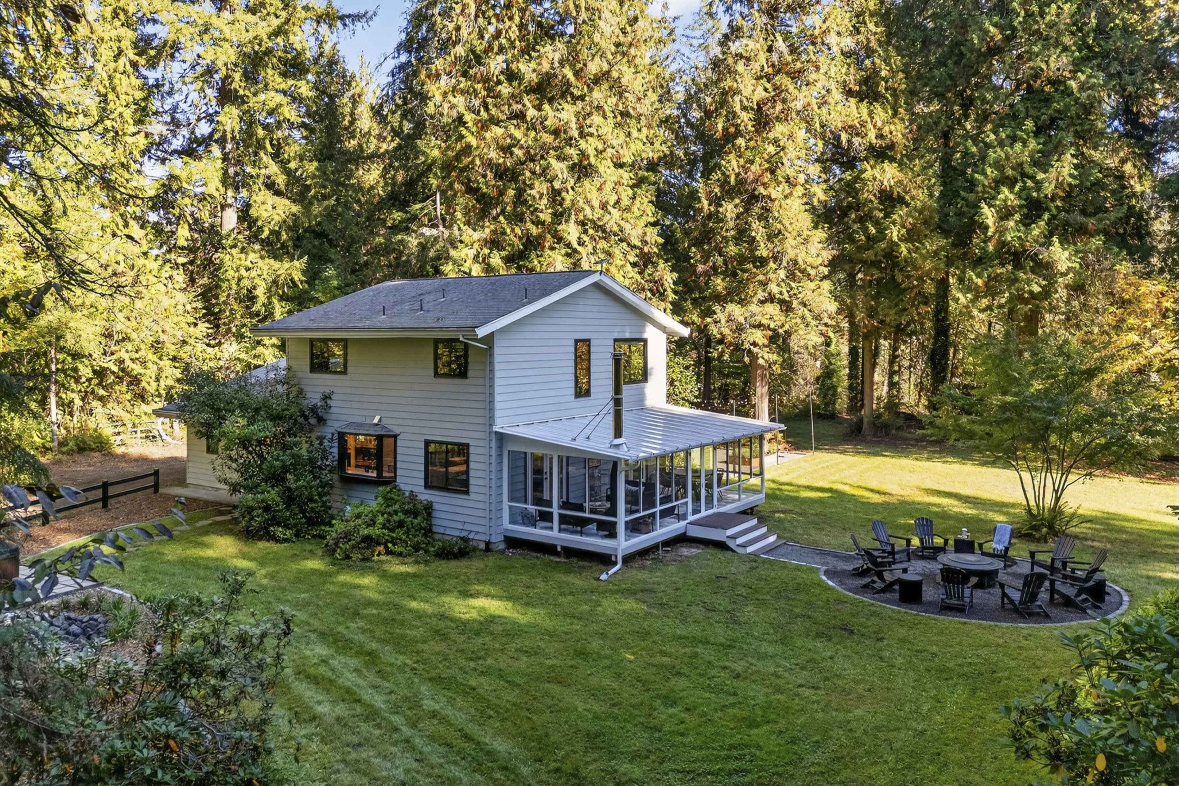A two-story house with a large deck is surrounded by tall trees in a grassy area.