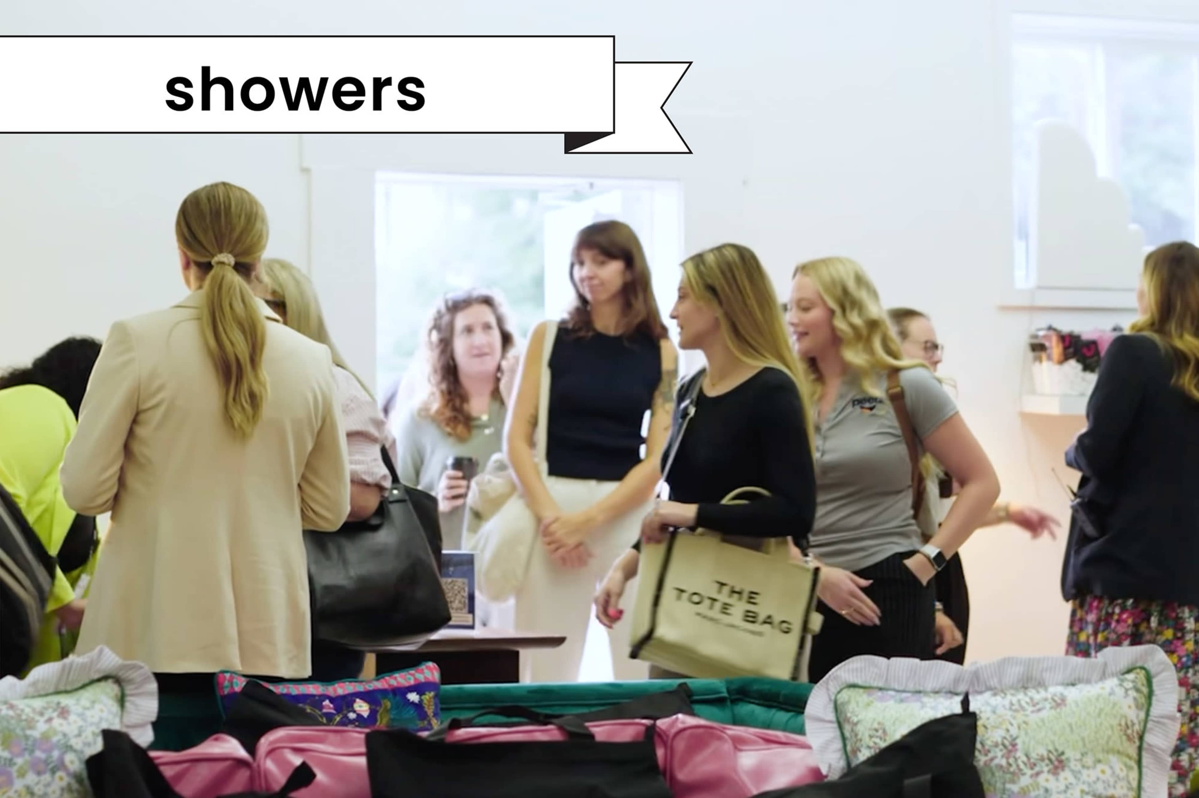 A group of people is gathered in a bright indoor space, mingling and holding bags while some browse items on tables.