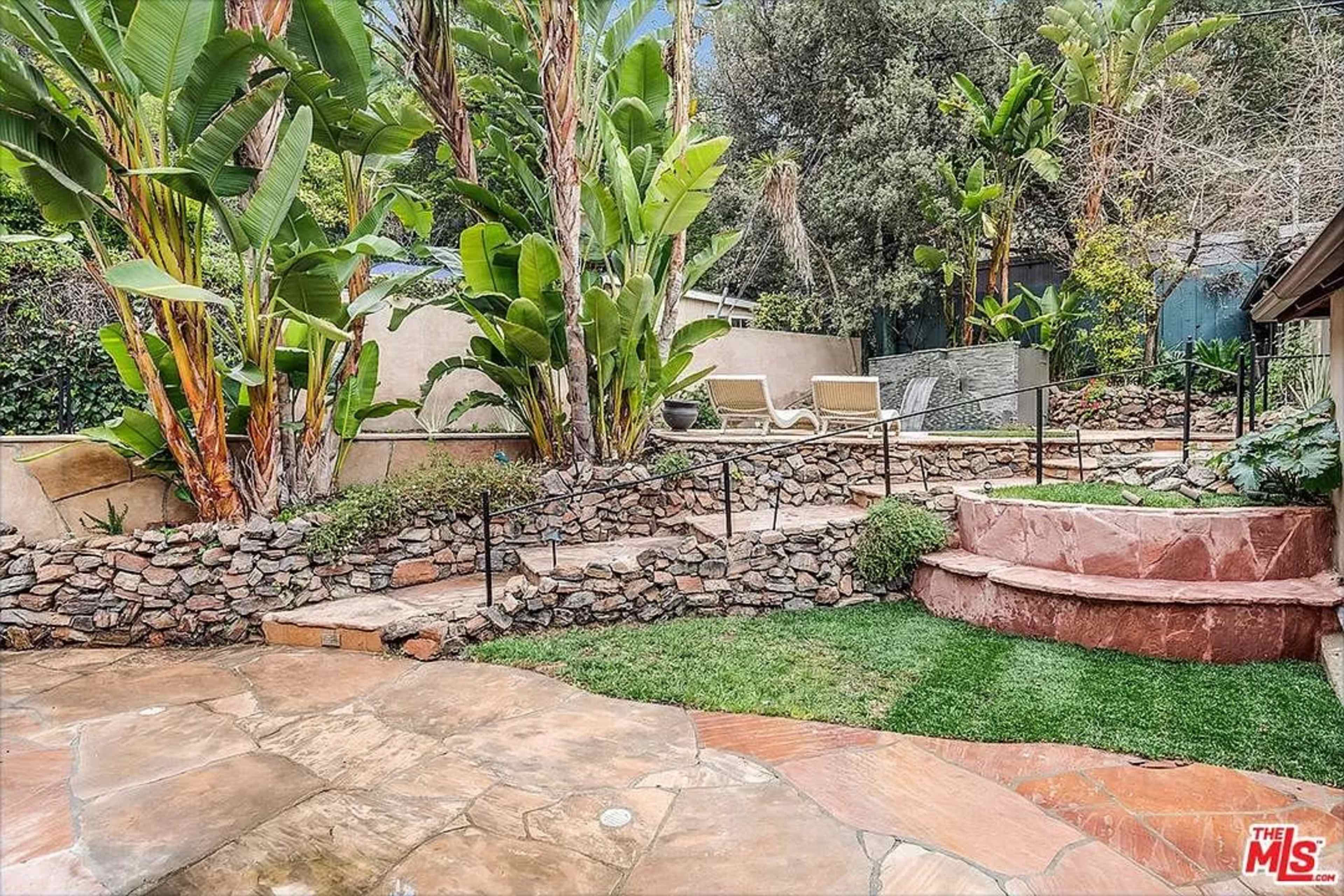 A landscaped backyard featuring stone pathways, terraced sections with terracotta steps, and lush green plants surrounding a seating area.