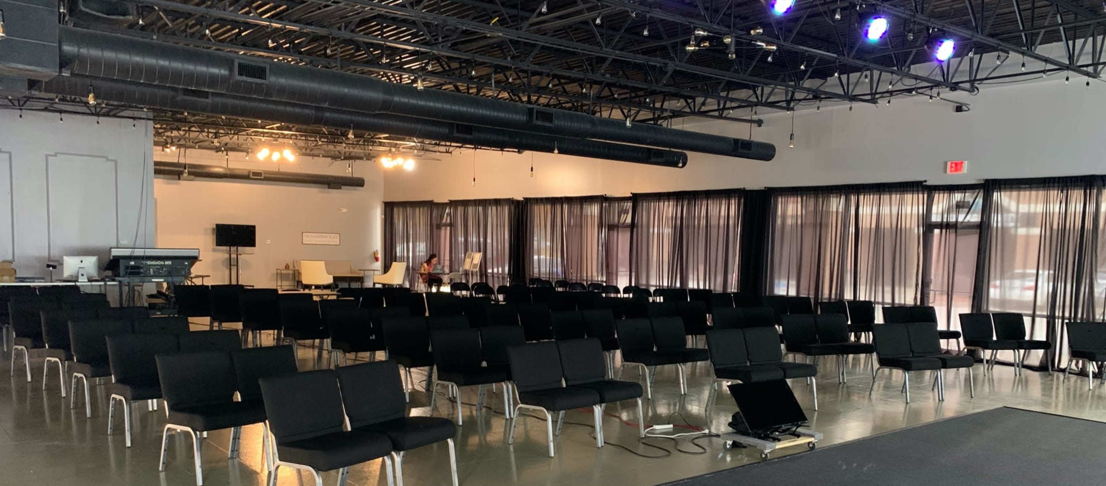 A spacious room set up with rows of black chairs facing a stage area, with large windows draped in black curtains.