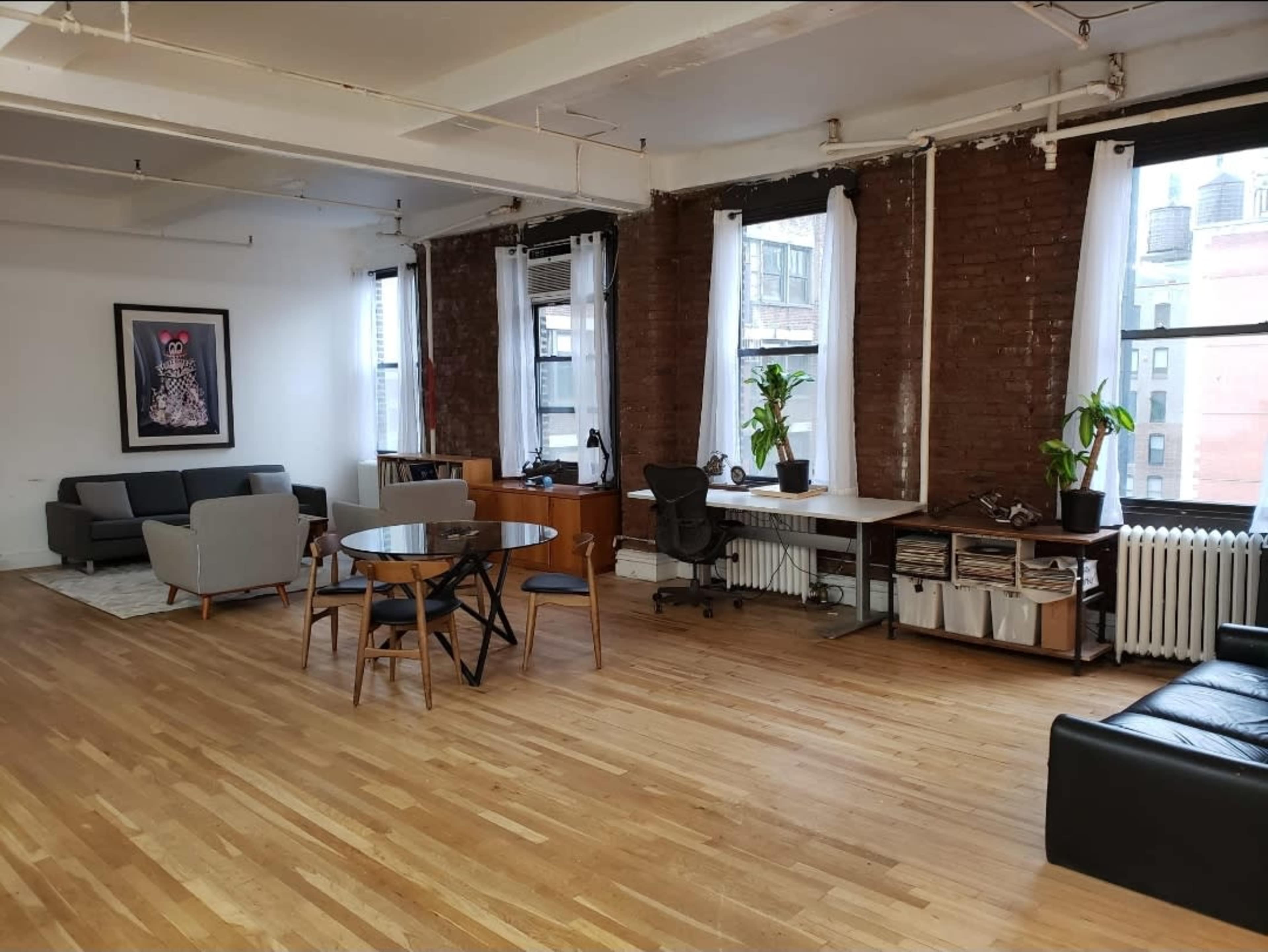 The image shows a spacious room with wooden flooring, featuring a sitting area with gray sofas, a dining table with chairs, and a desk against a brick wall with large windows.