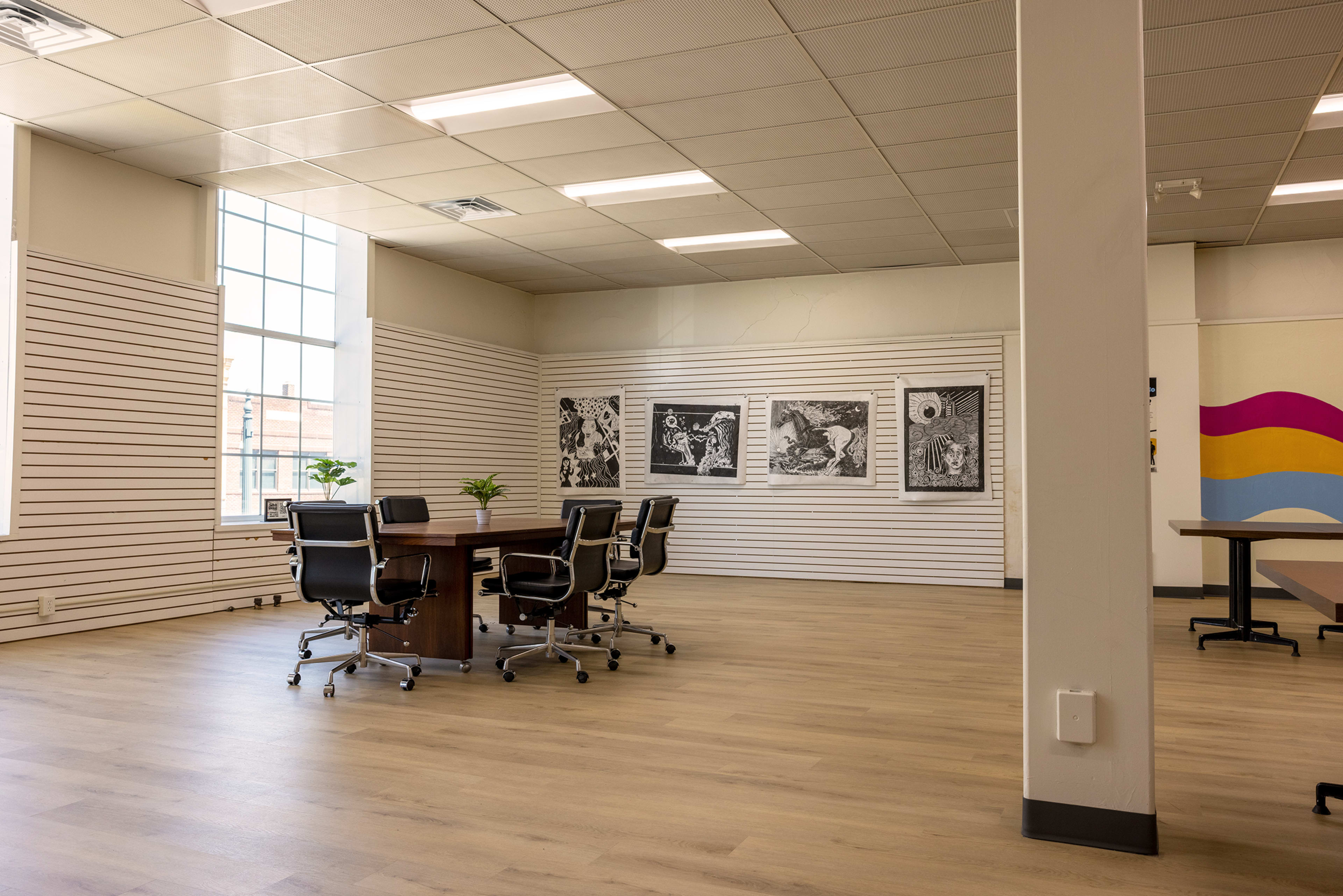 The image shows a spacious, minimalistic office with a large table surrounded by chairs and framed artwork on the walls.