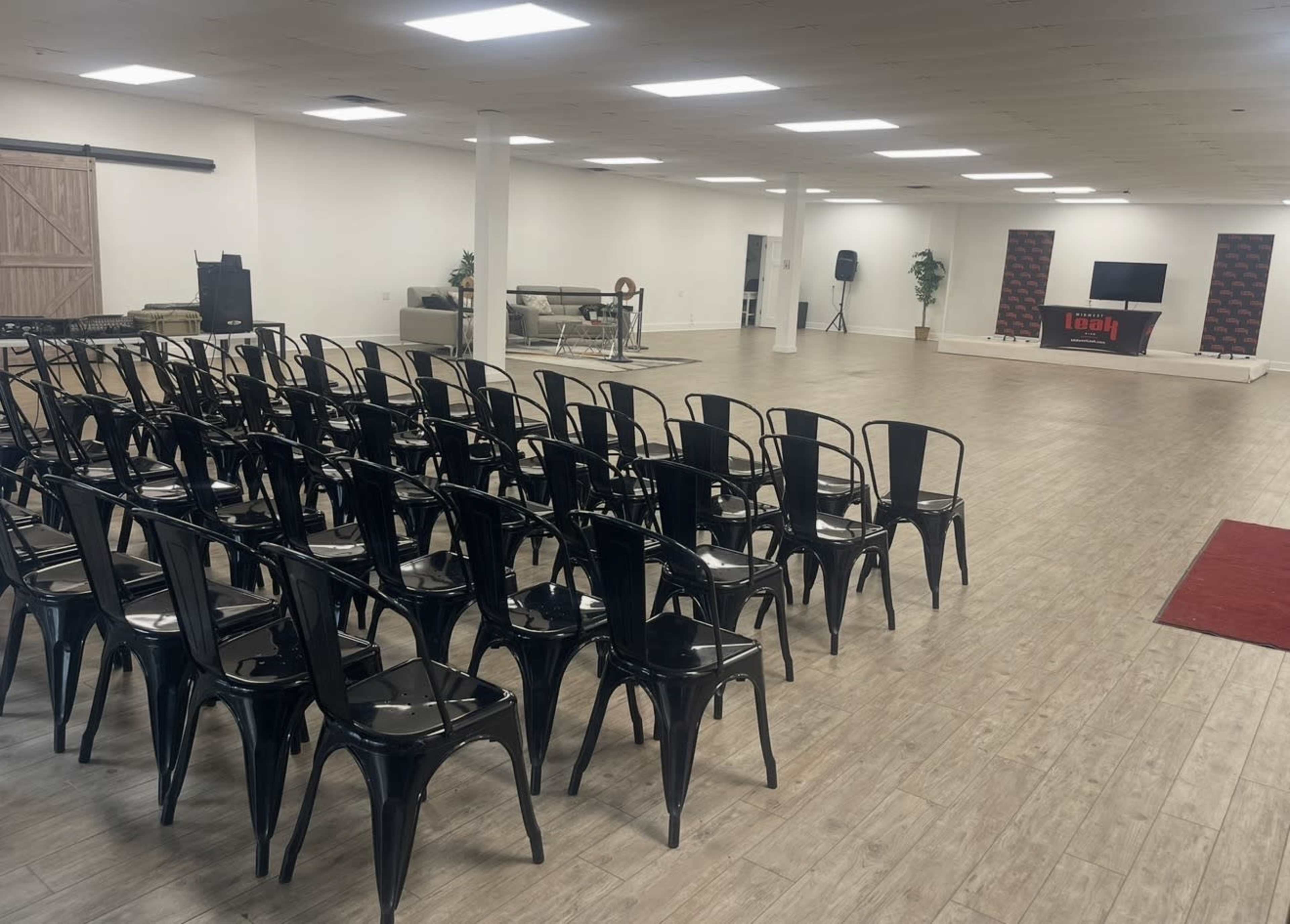 A spacious event room features rows of black chairs facing a raised platform with a TV and logo backdrop.