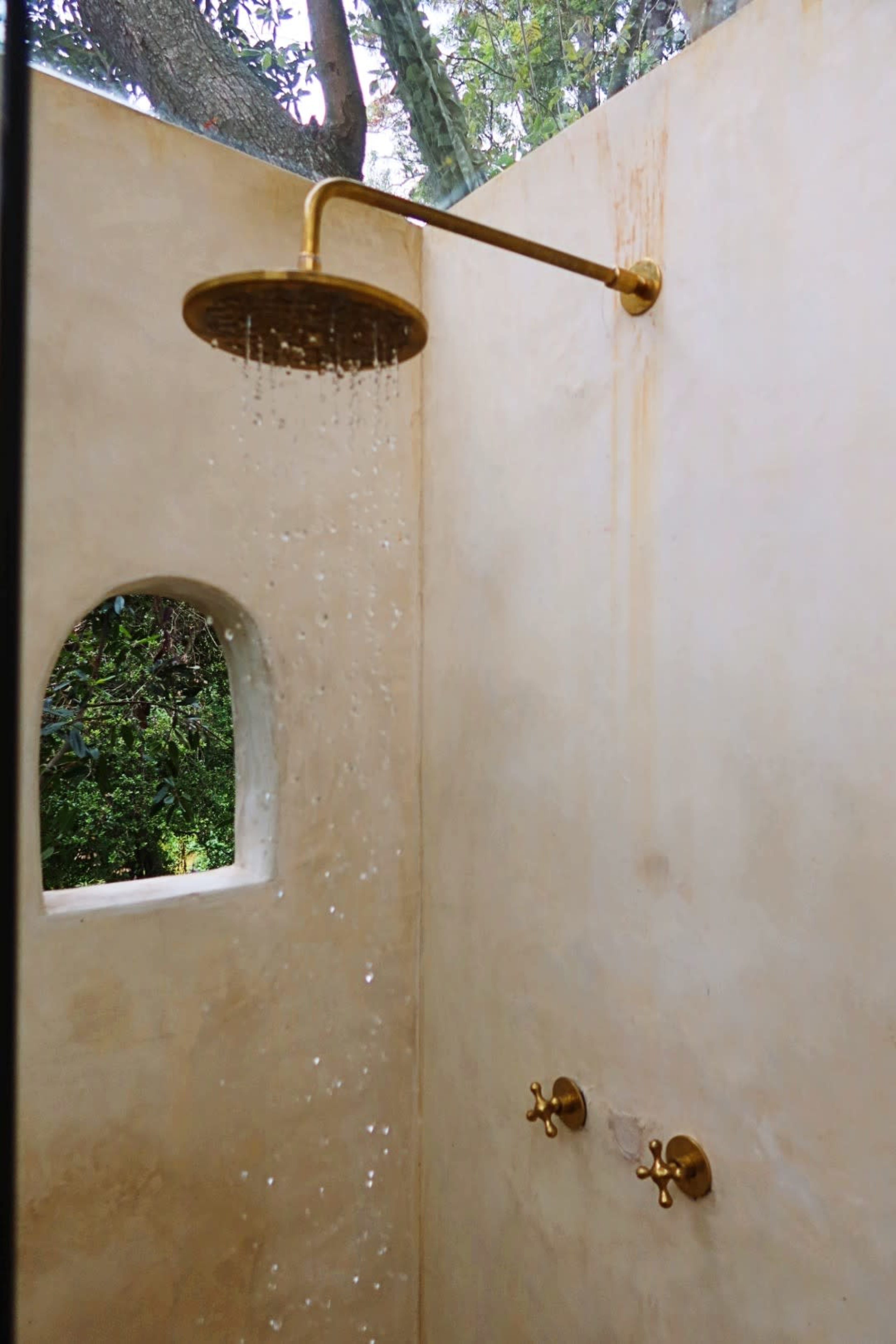 A gold showerhead streams water in an outdoor shower area with a smooth, light-colored wall and a small window.