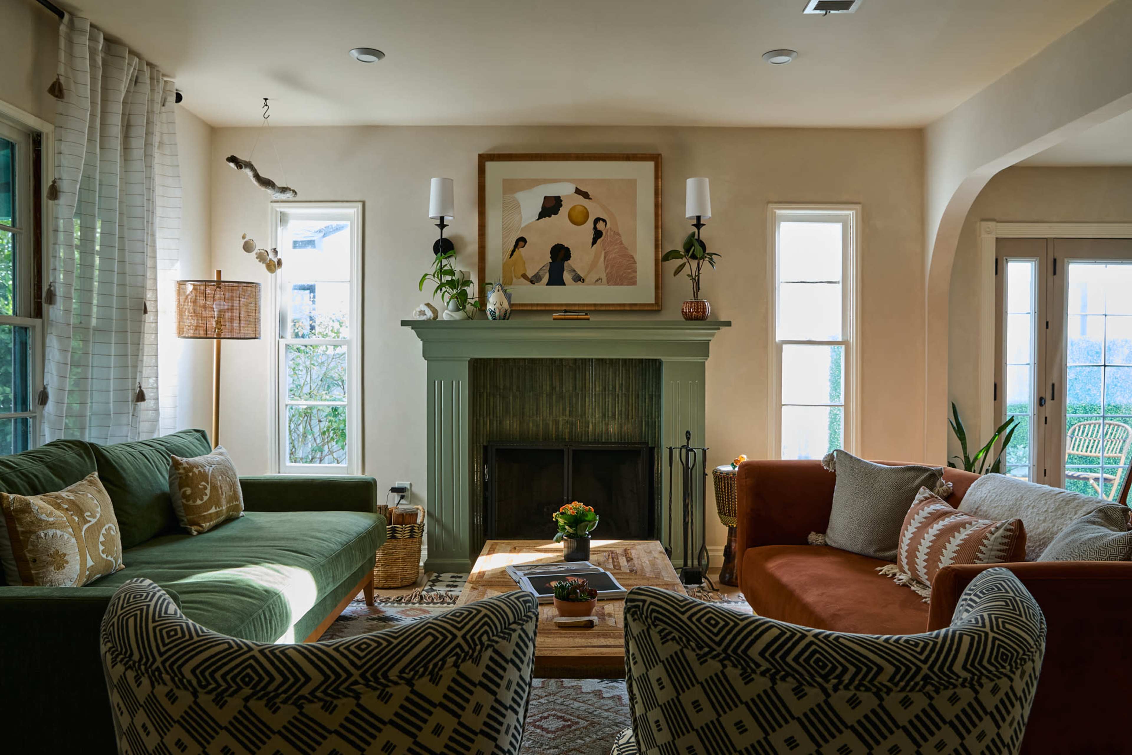 A cozy living room features a green couch, an orange armchair, and a decorative fireplace, surrounded by windows and plants.