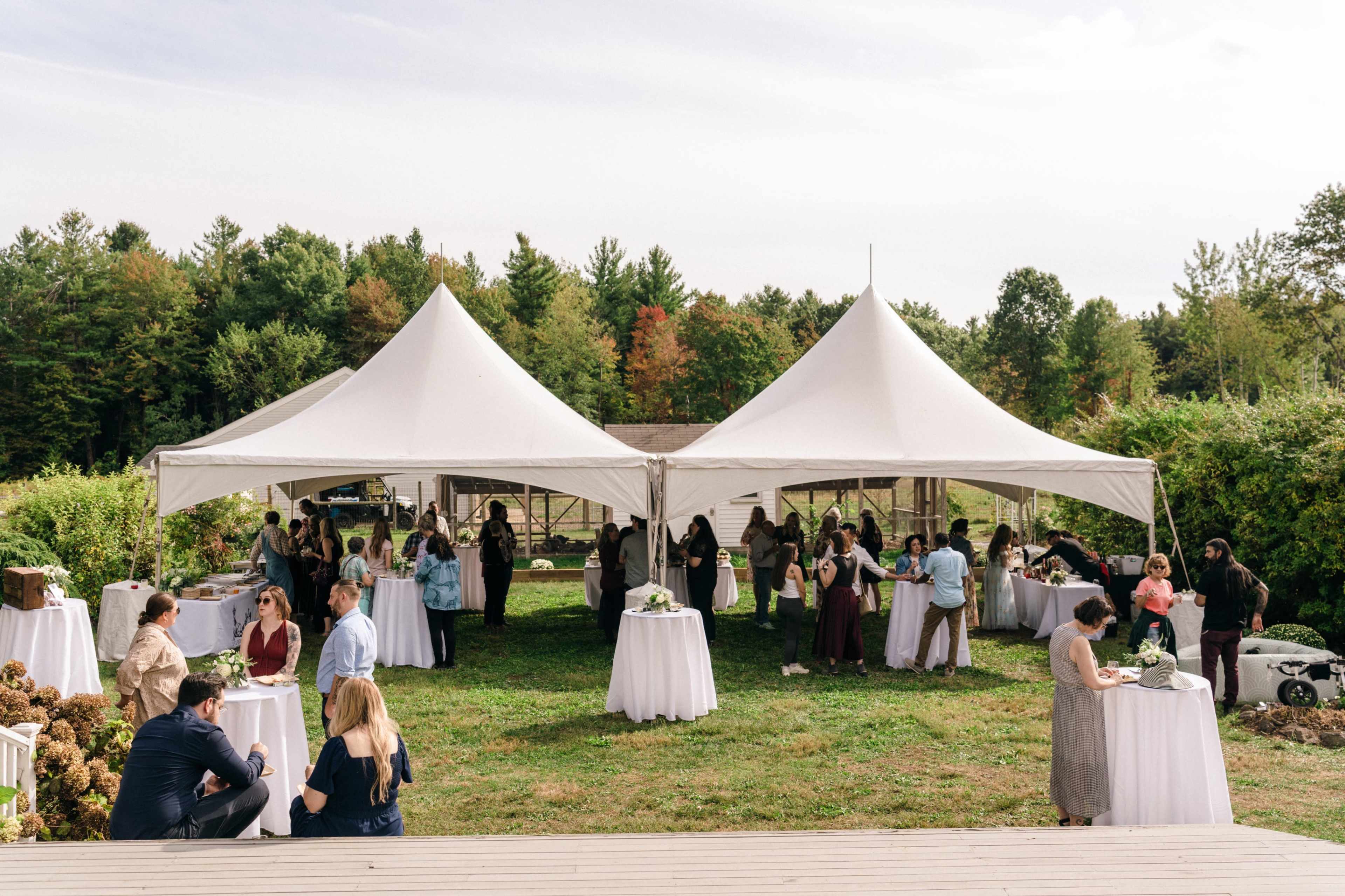A large outdoor event is taking place under two white tents, with numerous guests mingling and standing around tables on a grassy area.