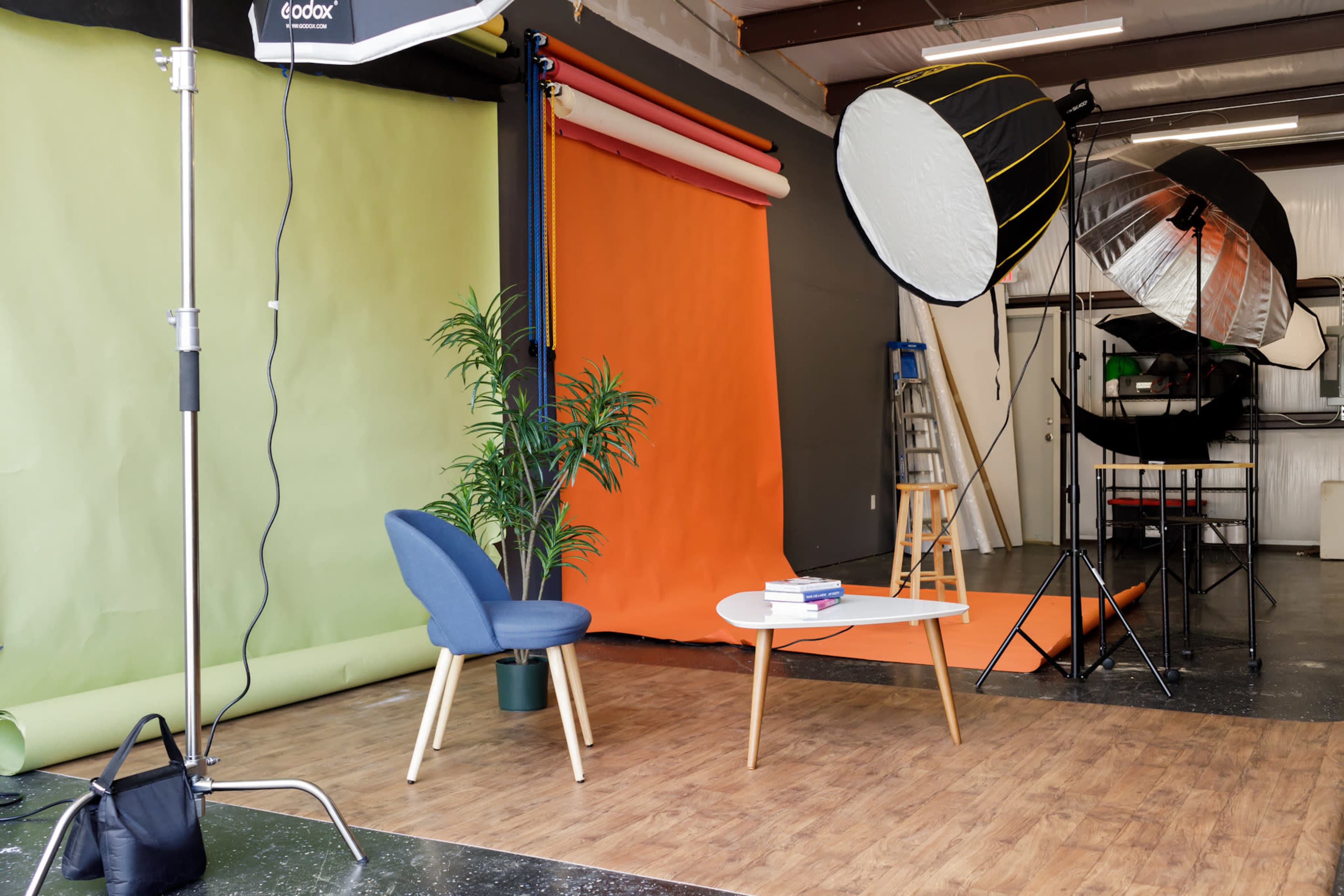 A photography studio features green and orange backdrops, a blue chair, a coffee table with books, and large softbox lights.