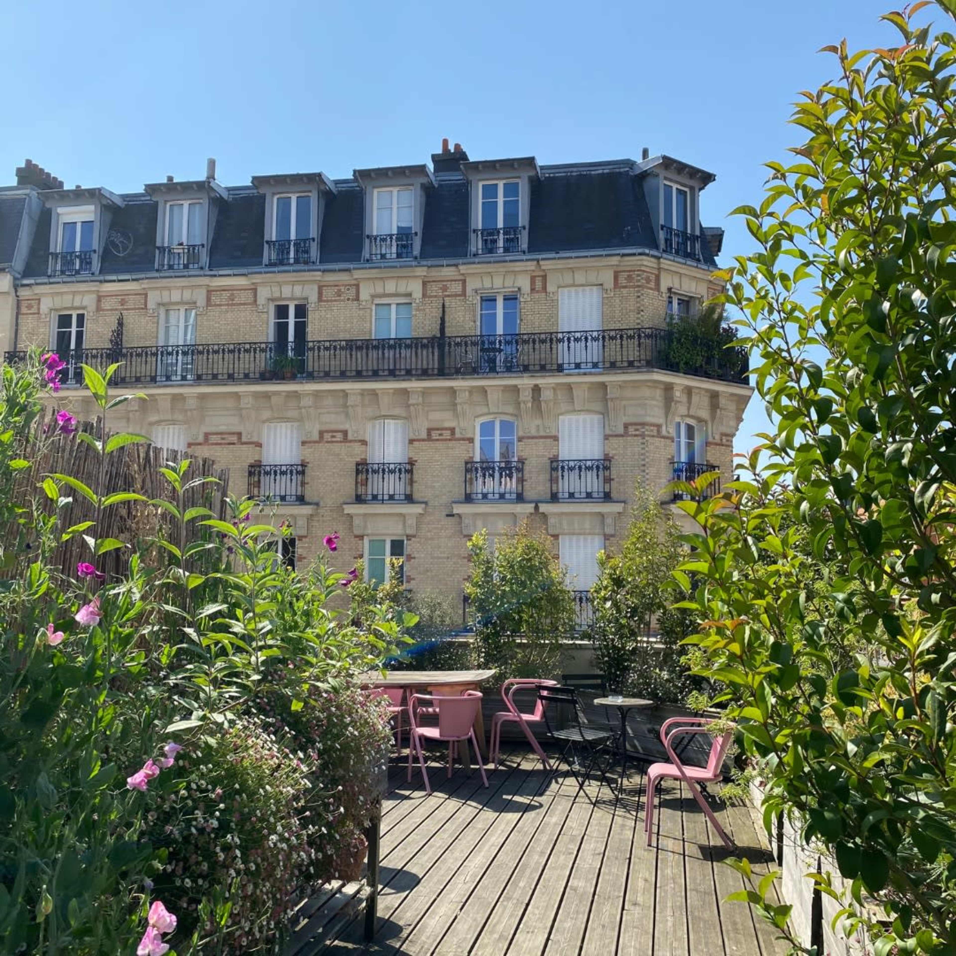 A brick building with balconies is seen from a terrace adorned with pink chairs and surrounded by greenery and flowers.