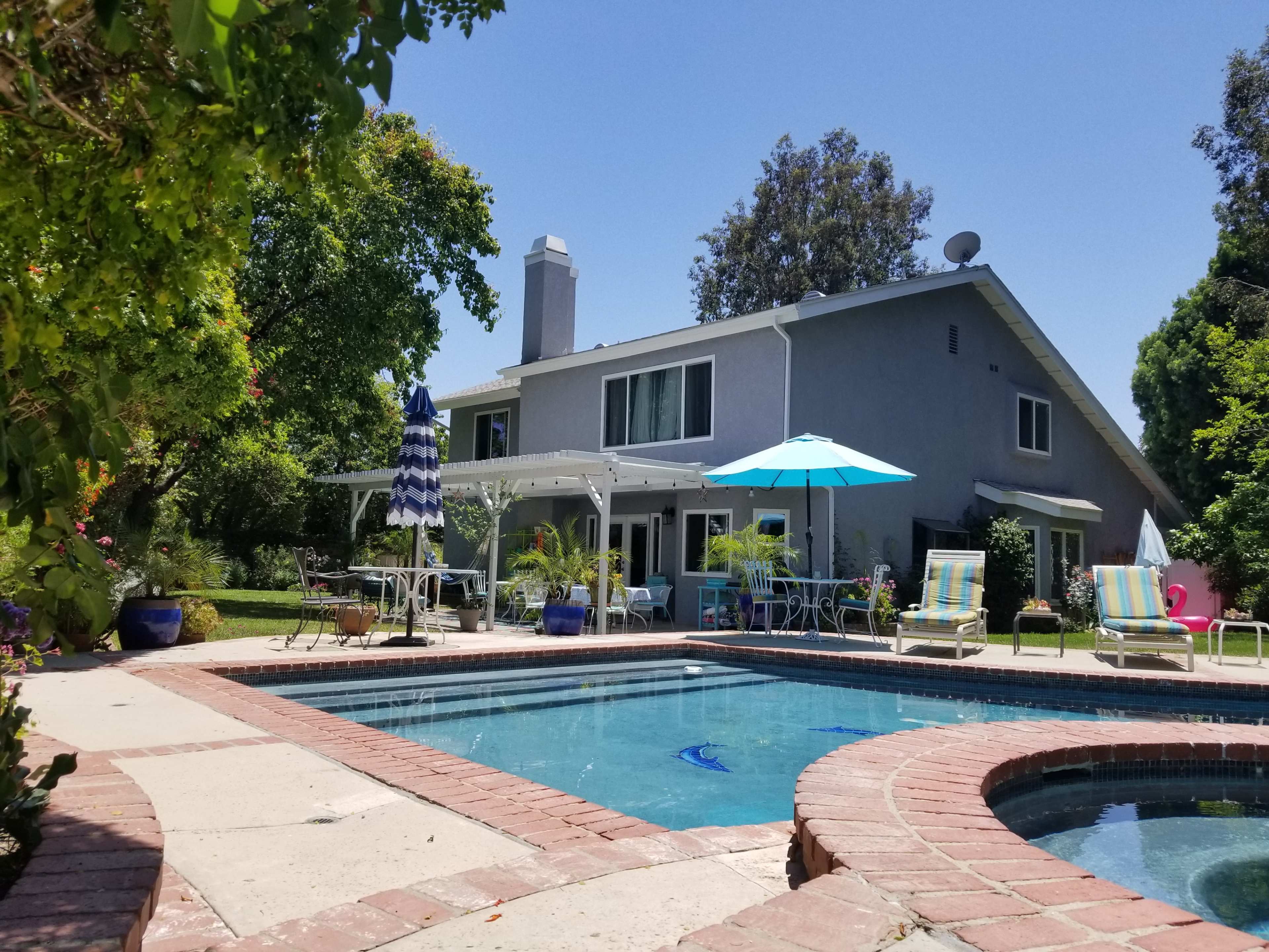 A two-story house with a swimming pool and patio furniture is surrounded by greenery and a clear blue sky.
