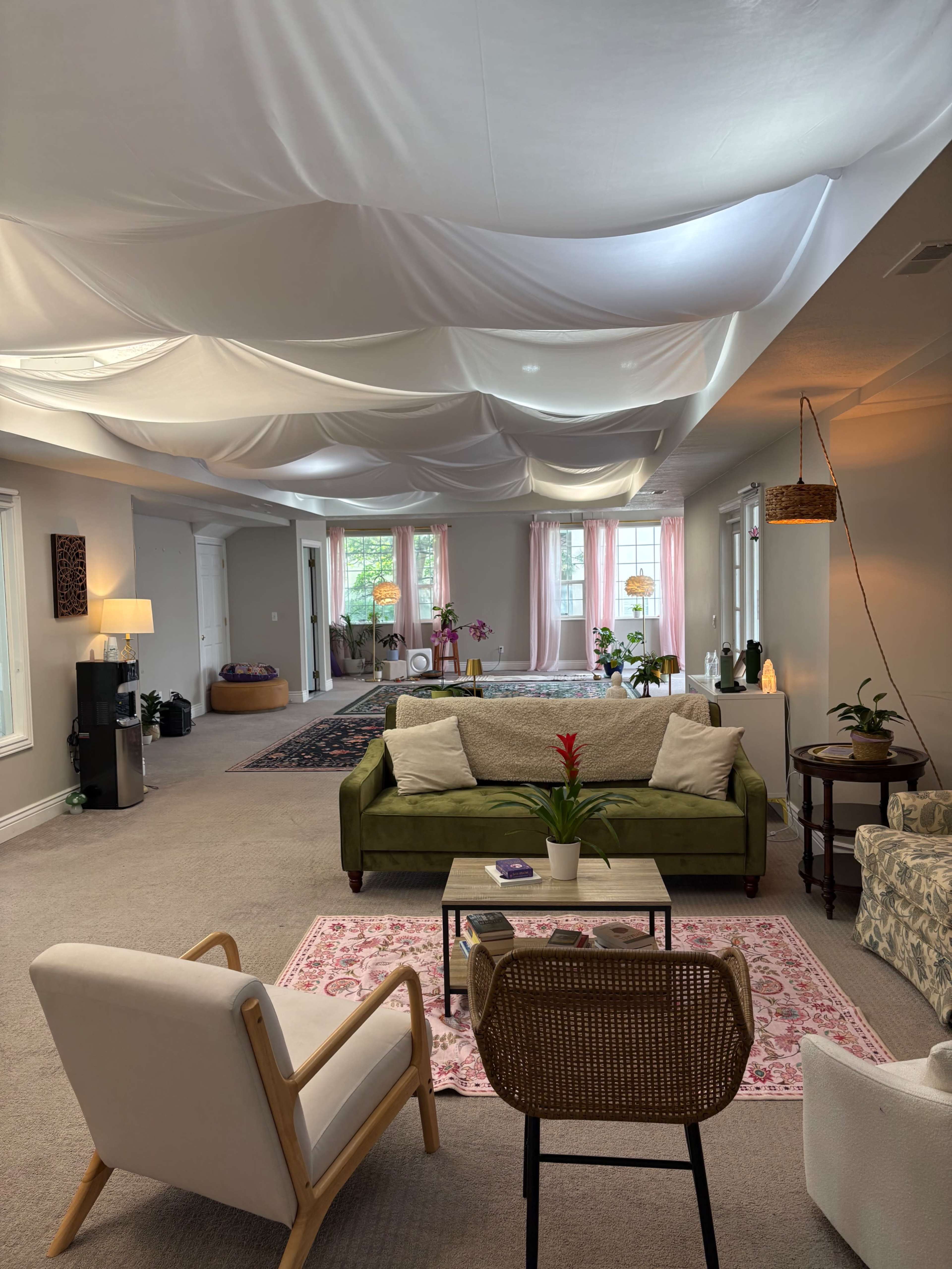 A spacious room features a green sofa, various plants, and draped fabric on the ceiling, creating a soft, inviting atmosphere.