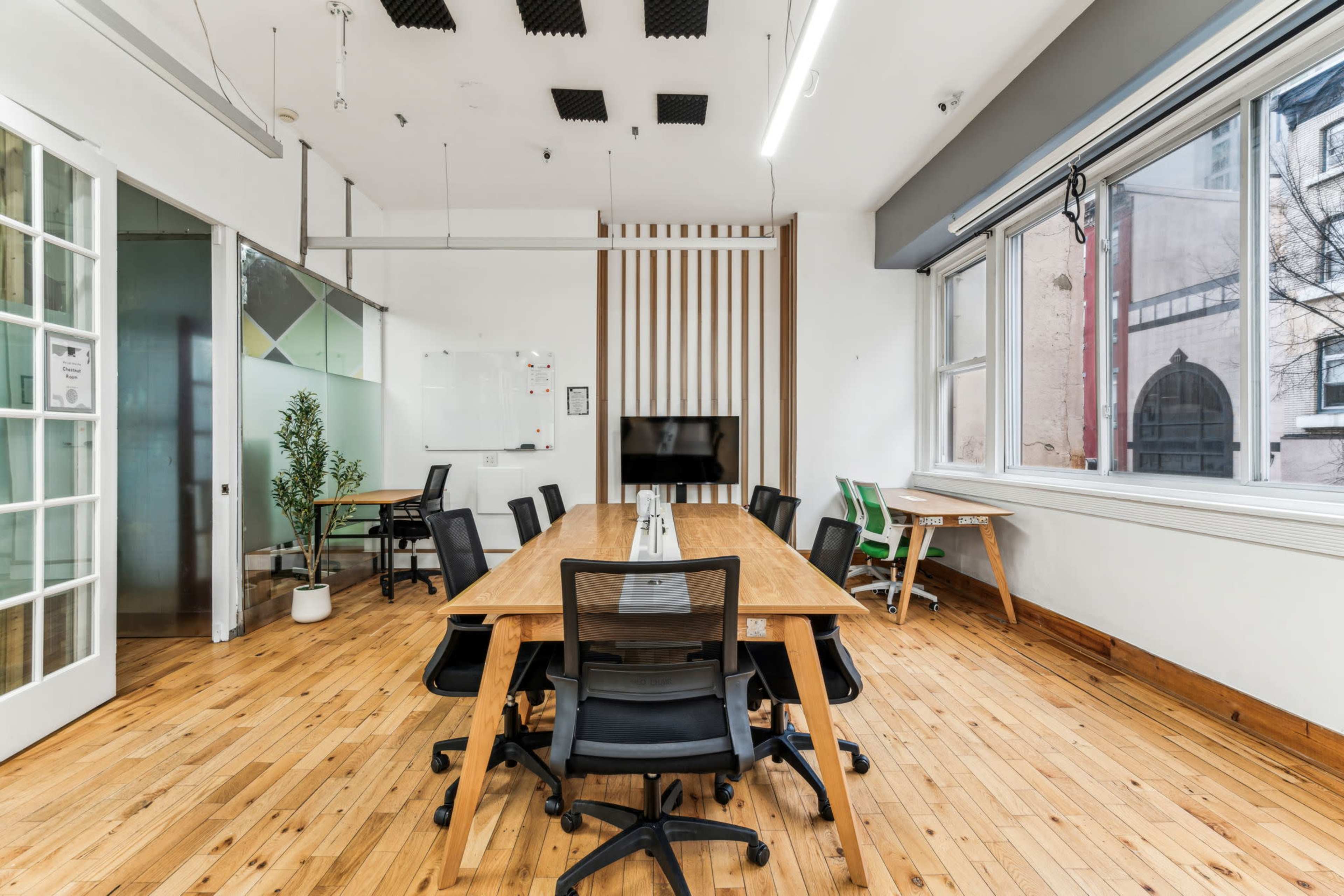 The image shows a bright, modern meeting room with a wooden table, black chairs, and large windows.