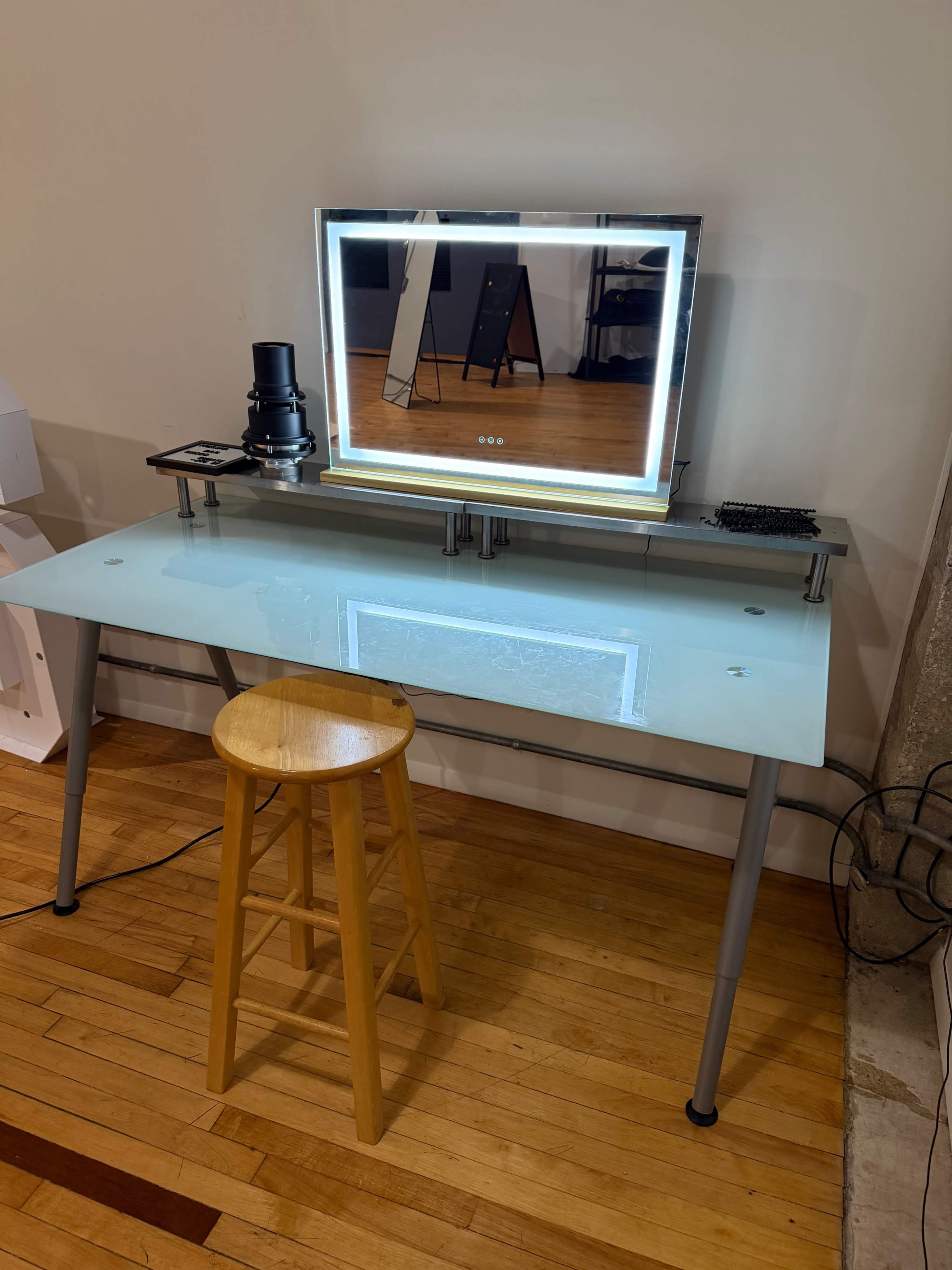 The image shows a modern glass-top desk with a stool, a mirror with LED lighting, and a small black device on its surface.