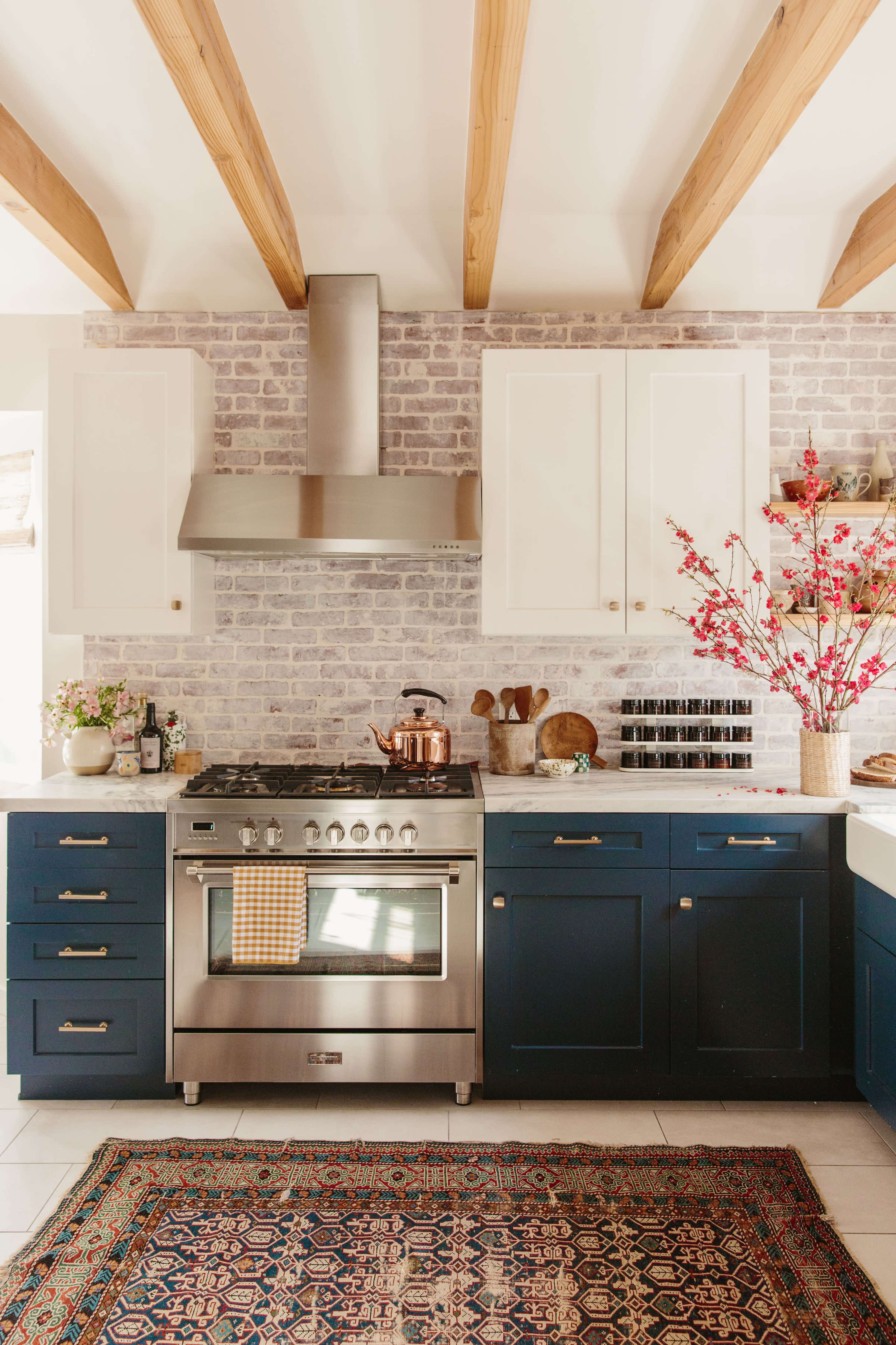A modern kitchen features blue cabinets, a stainless steel stove, and a patterned rug, with exposed wooden beams and decorative elements on the counters.