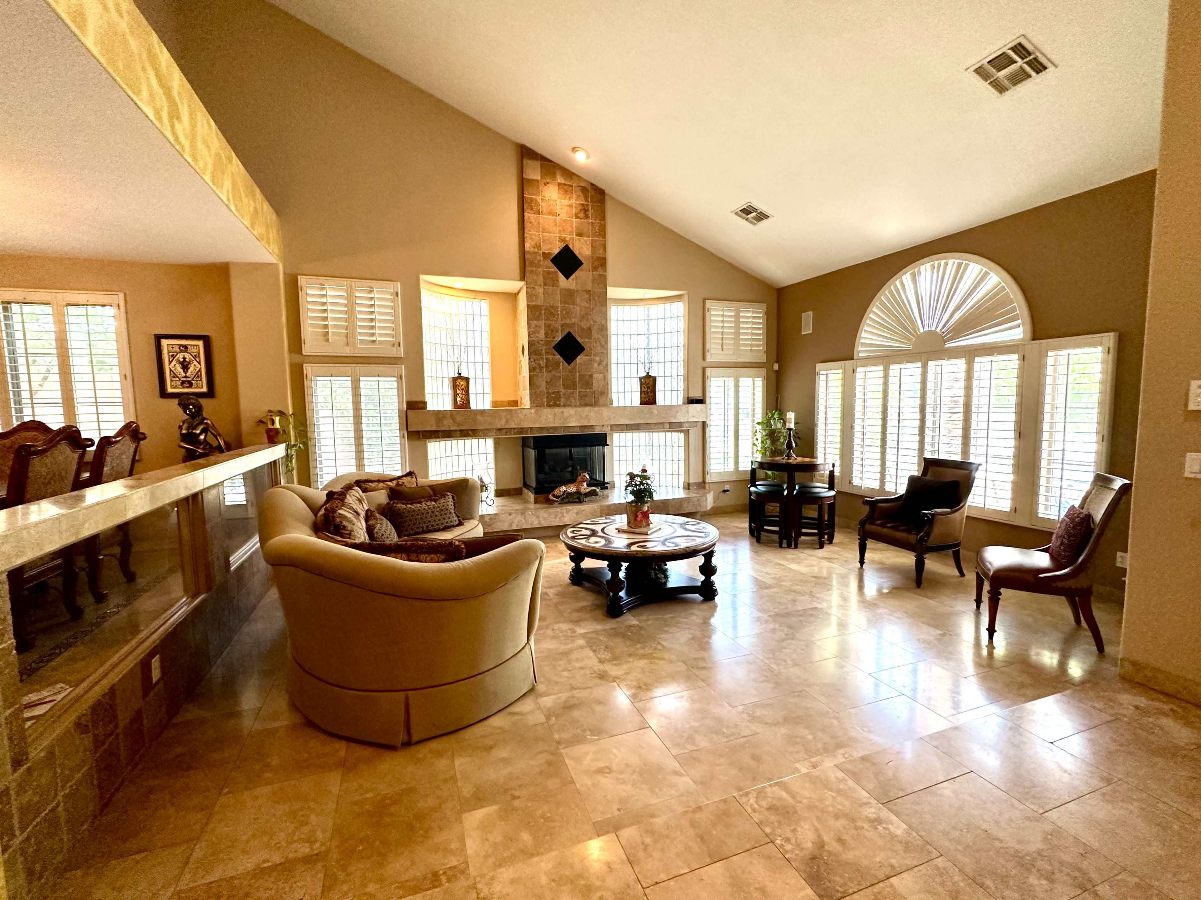 A spacious living room features a central fireplace, large windows with shutters, and a mix of comfortable seating around a round coffee table.