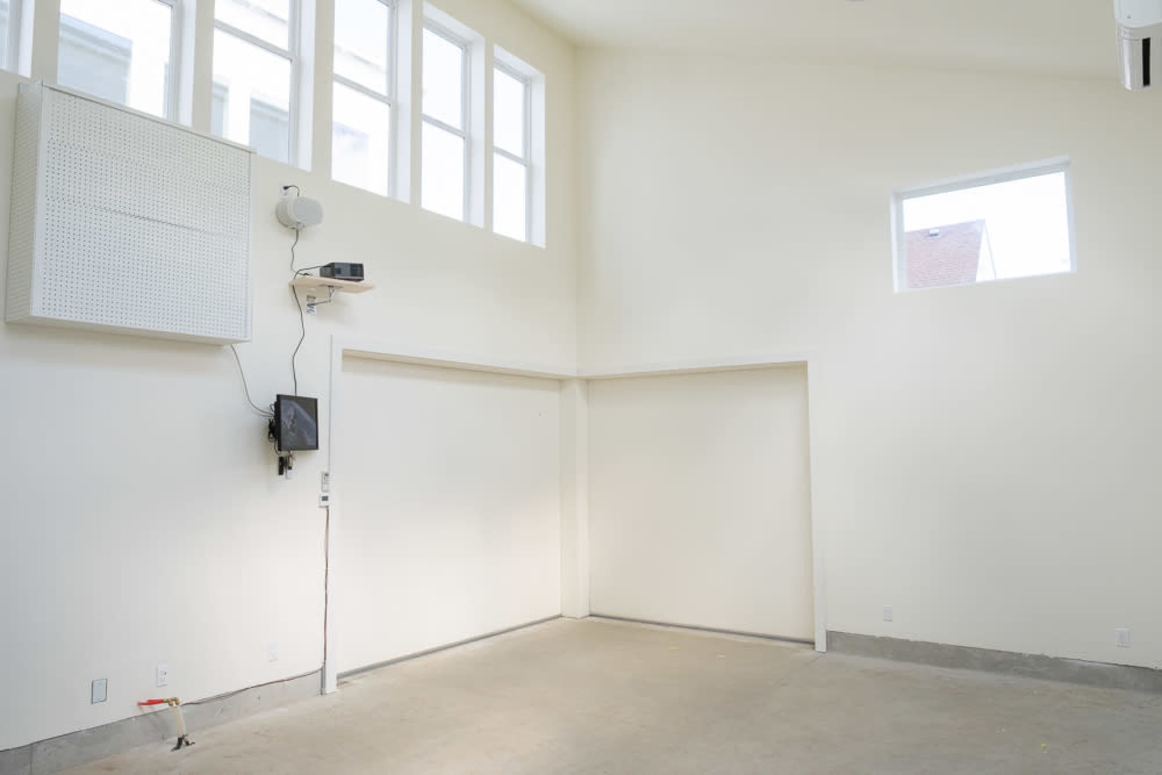 The image shows a vacant interior space with white walls, high ceilings, and one window on each side, along with a mounted projector and a small screen on the wall.