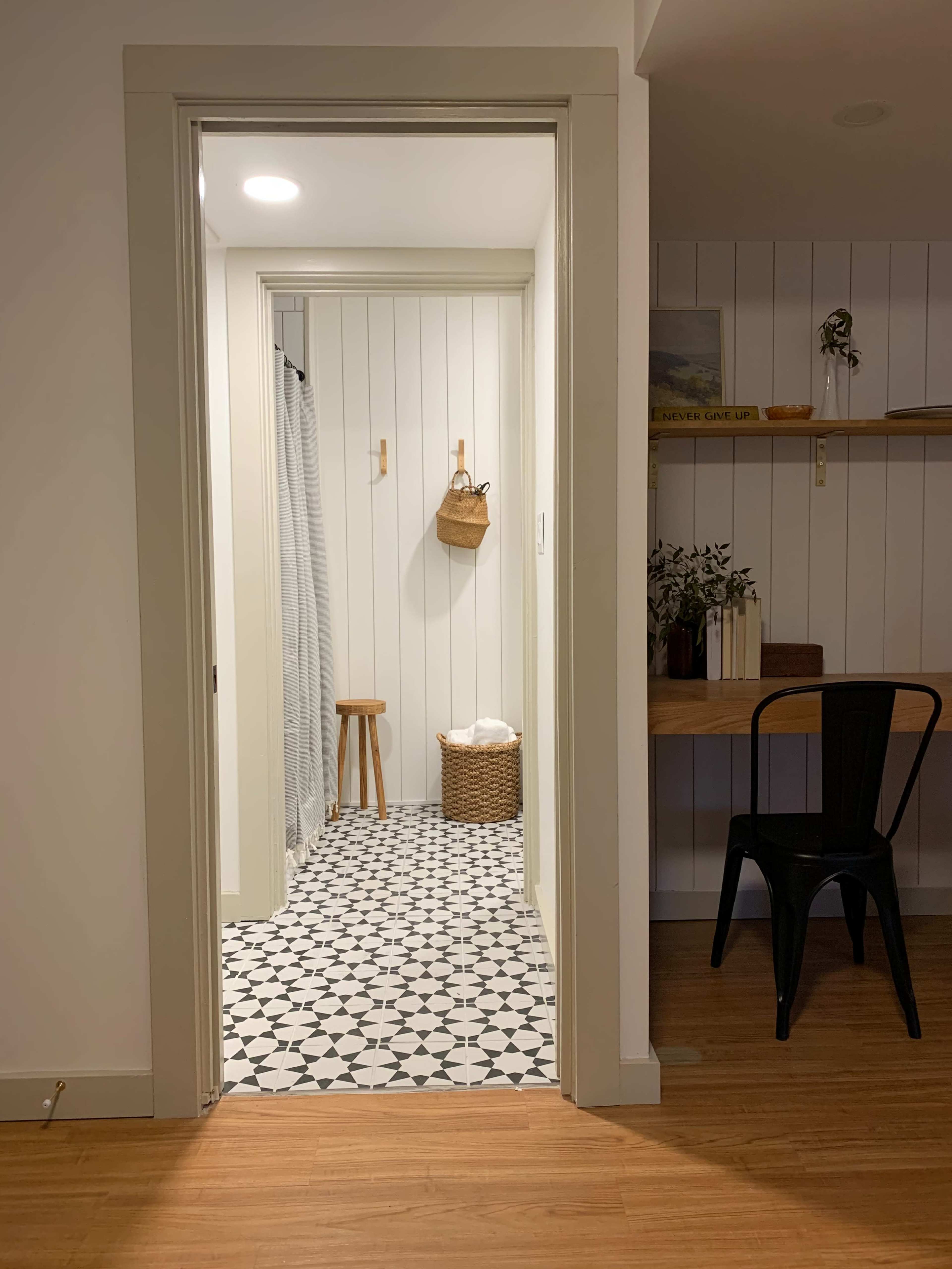 A bathroom entrance with black and white patterned tiles, a wooden stool, a basket, and a hanging shower curtain.