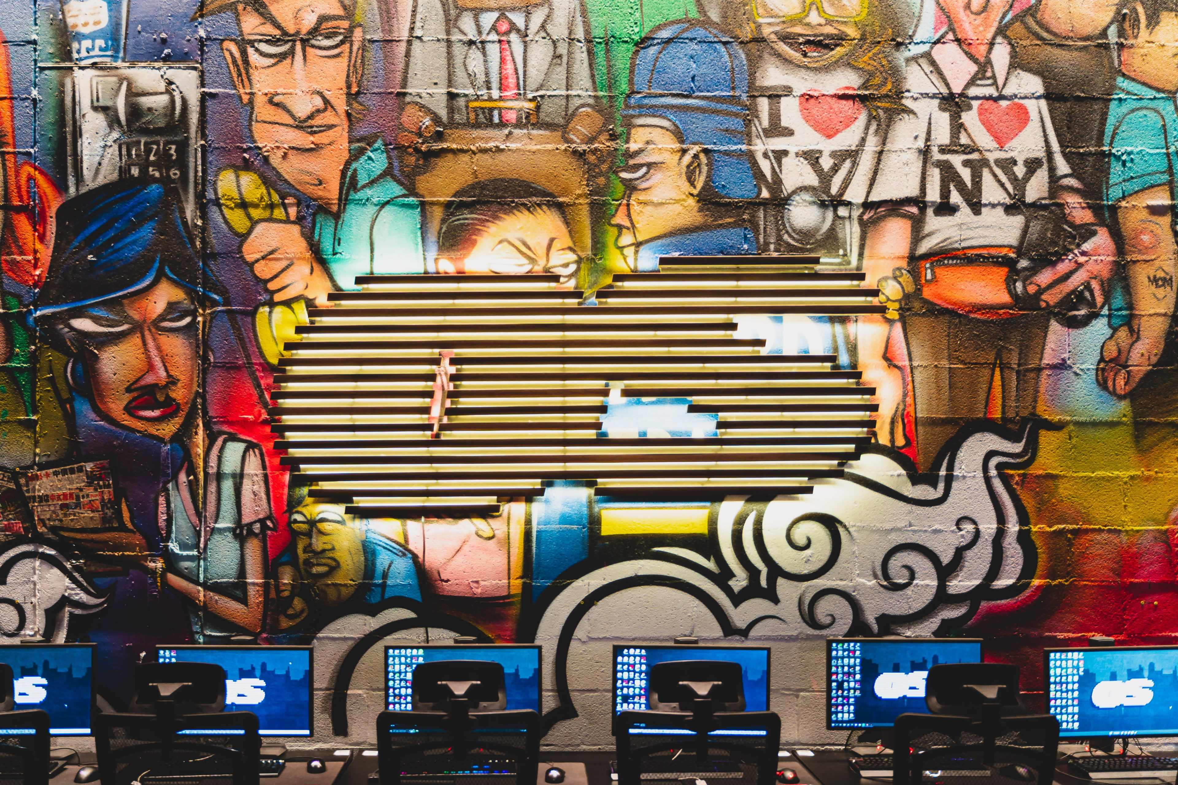 A vibrant graffiti wall with a prominent logo, surrounded by various colorful characters, and several computer monitors positioned in front of it.