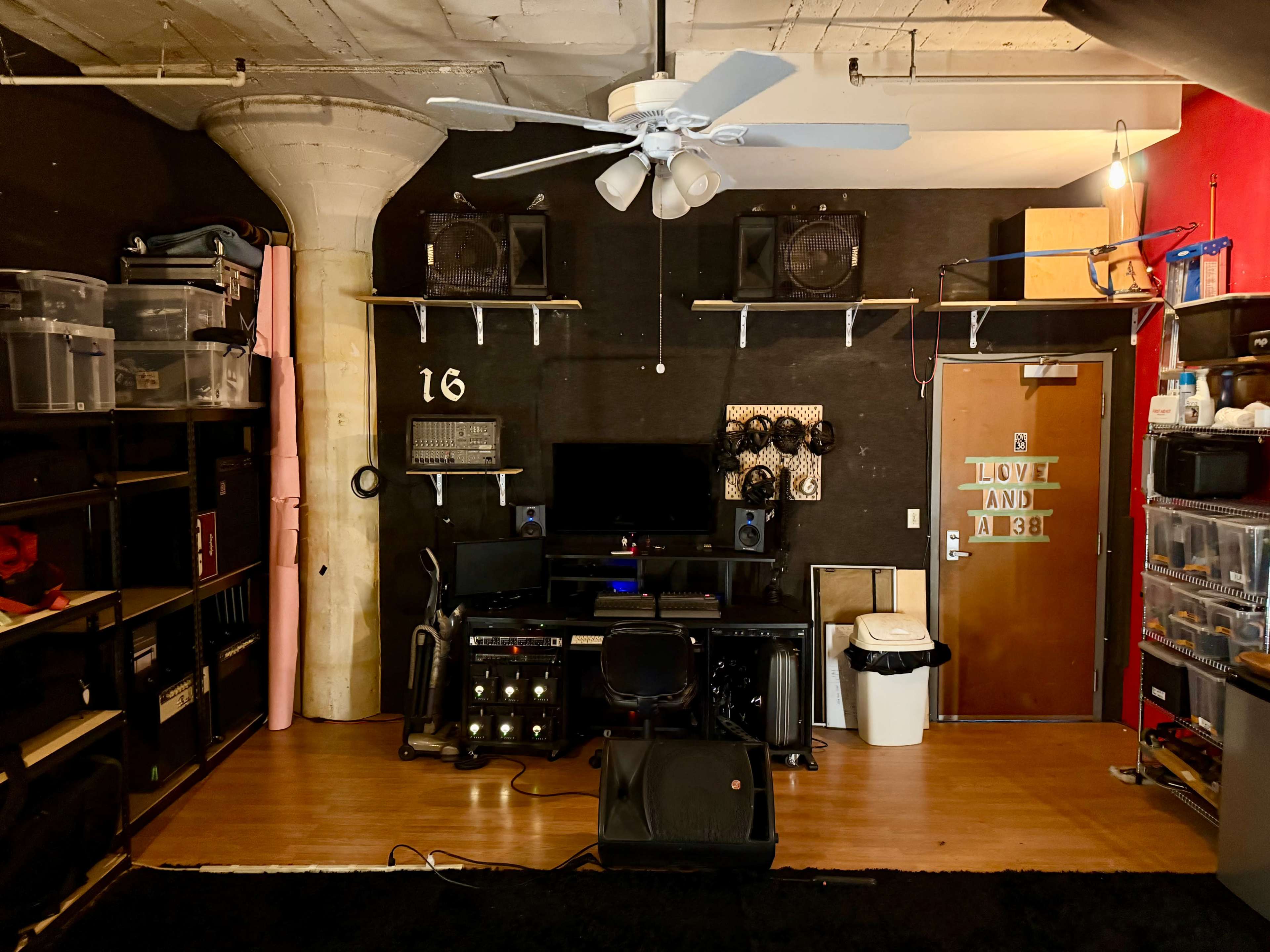 The image shows a organized studio space with a black wall, computer setup, shelving units, and a door that features a "Love" and "AB" sign.