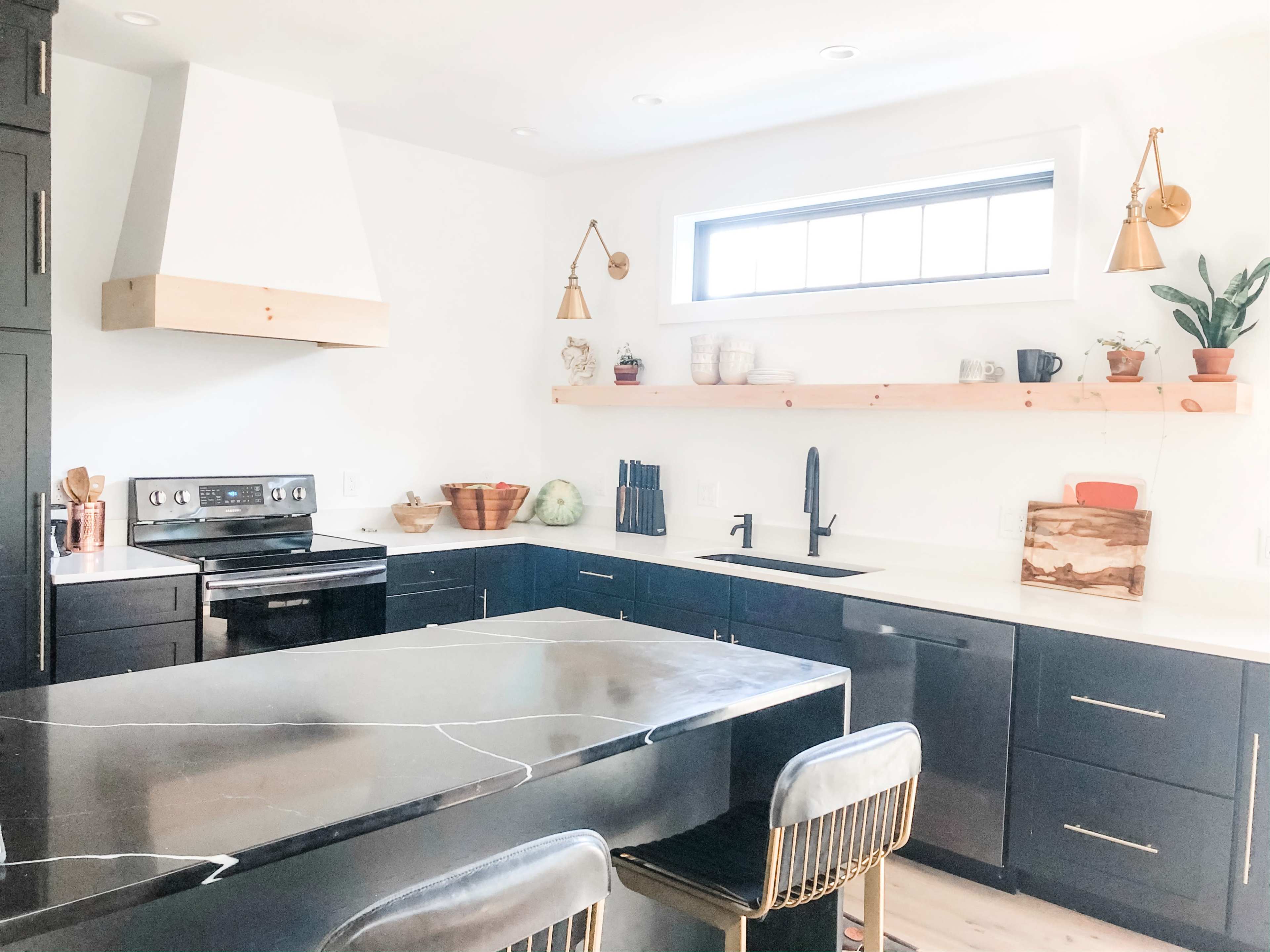 A modern kitchen features black cabinets, a central island with a black marble countertop, and wooden shelving above a stainless steel sink.