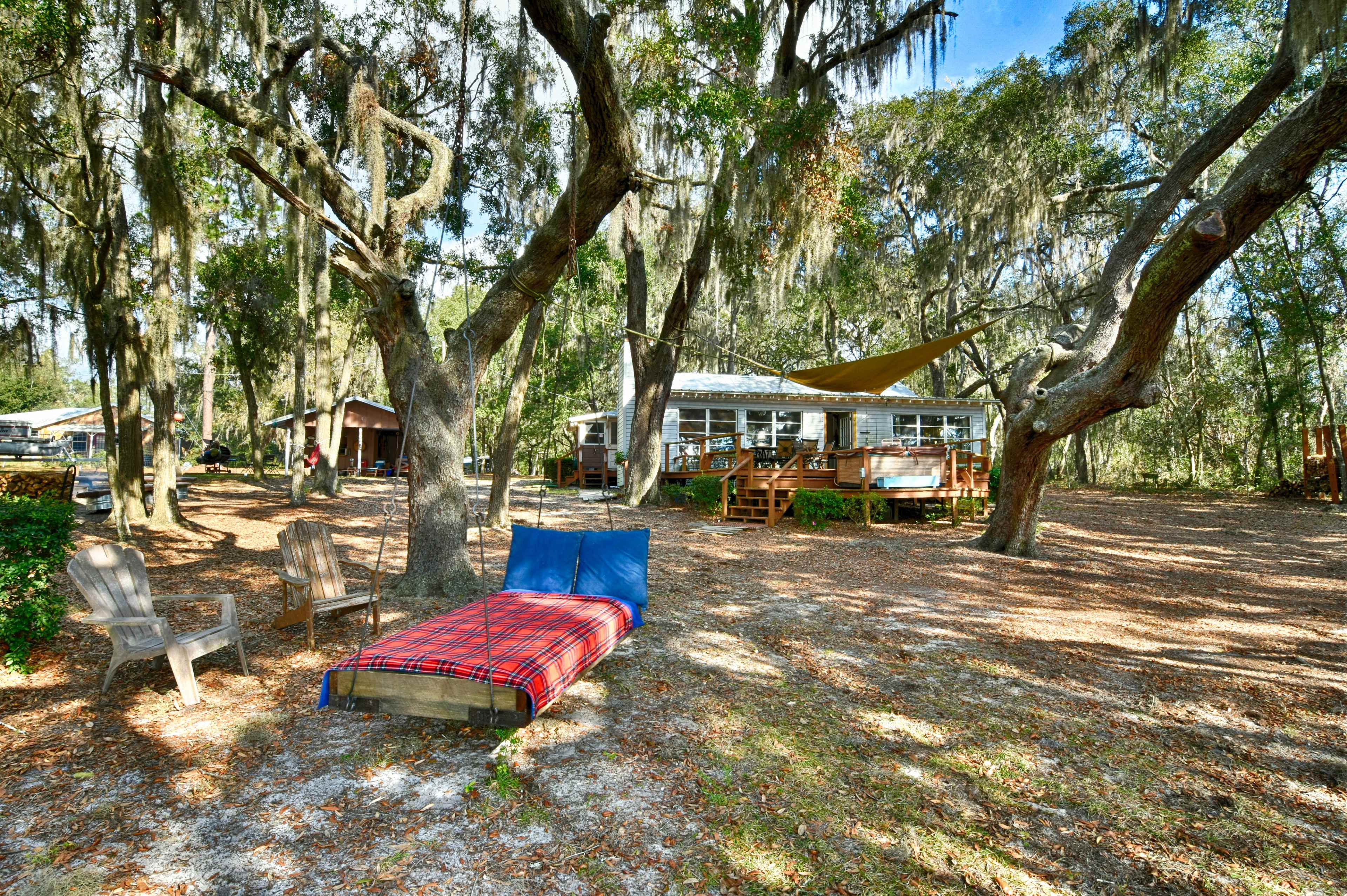 A wooden bed with a blue mattress is positioned in a grassy area near a house surrounded by trees with Spanish moss.