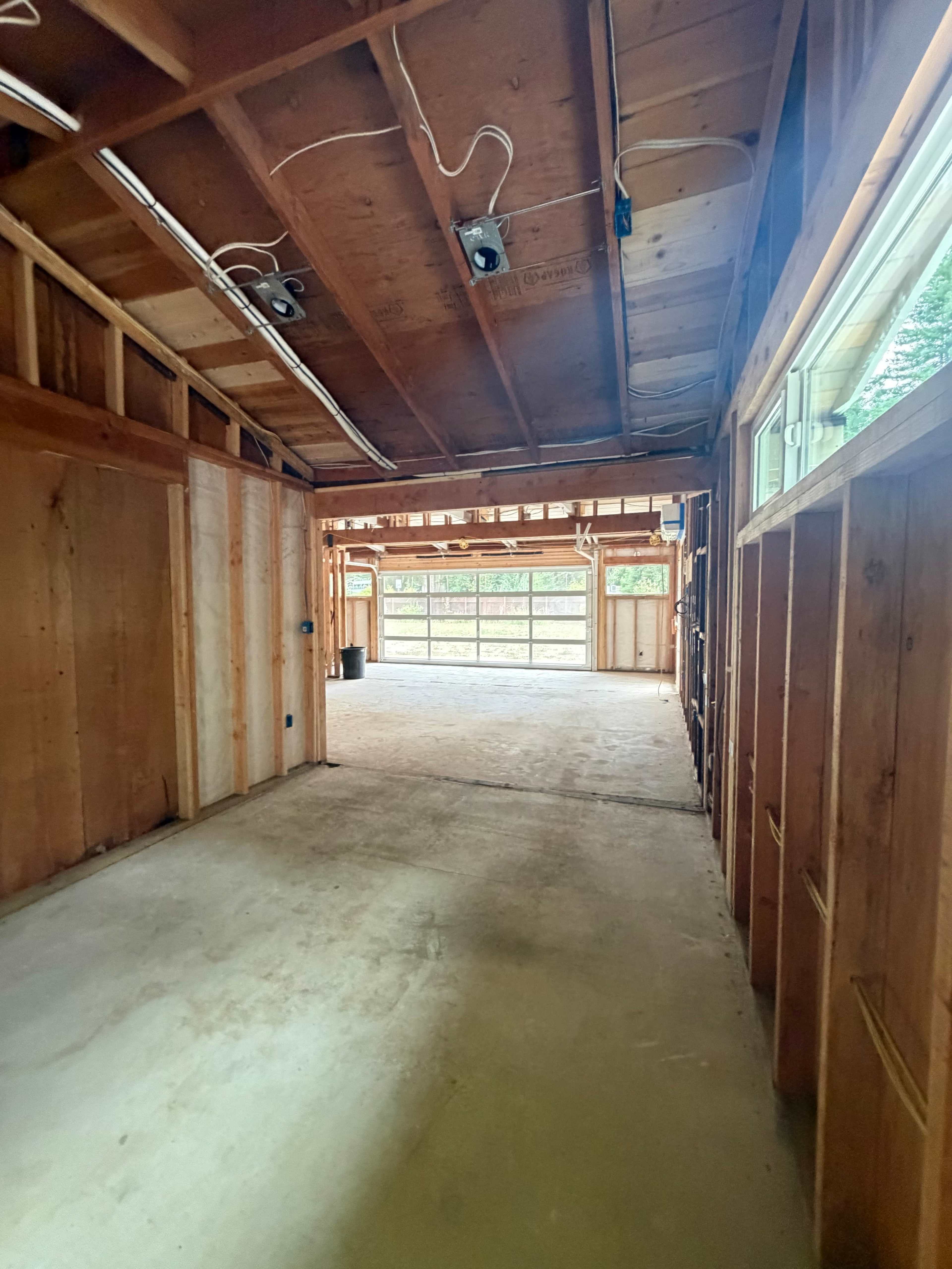 The image shows an empty, partially constructed interior space with exposed wooden beams and framing, leading to a large garage opening.