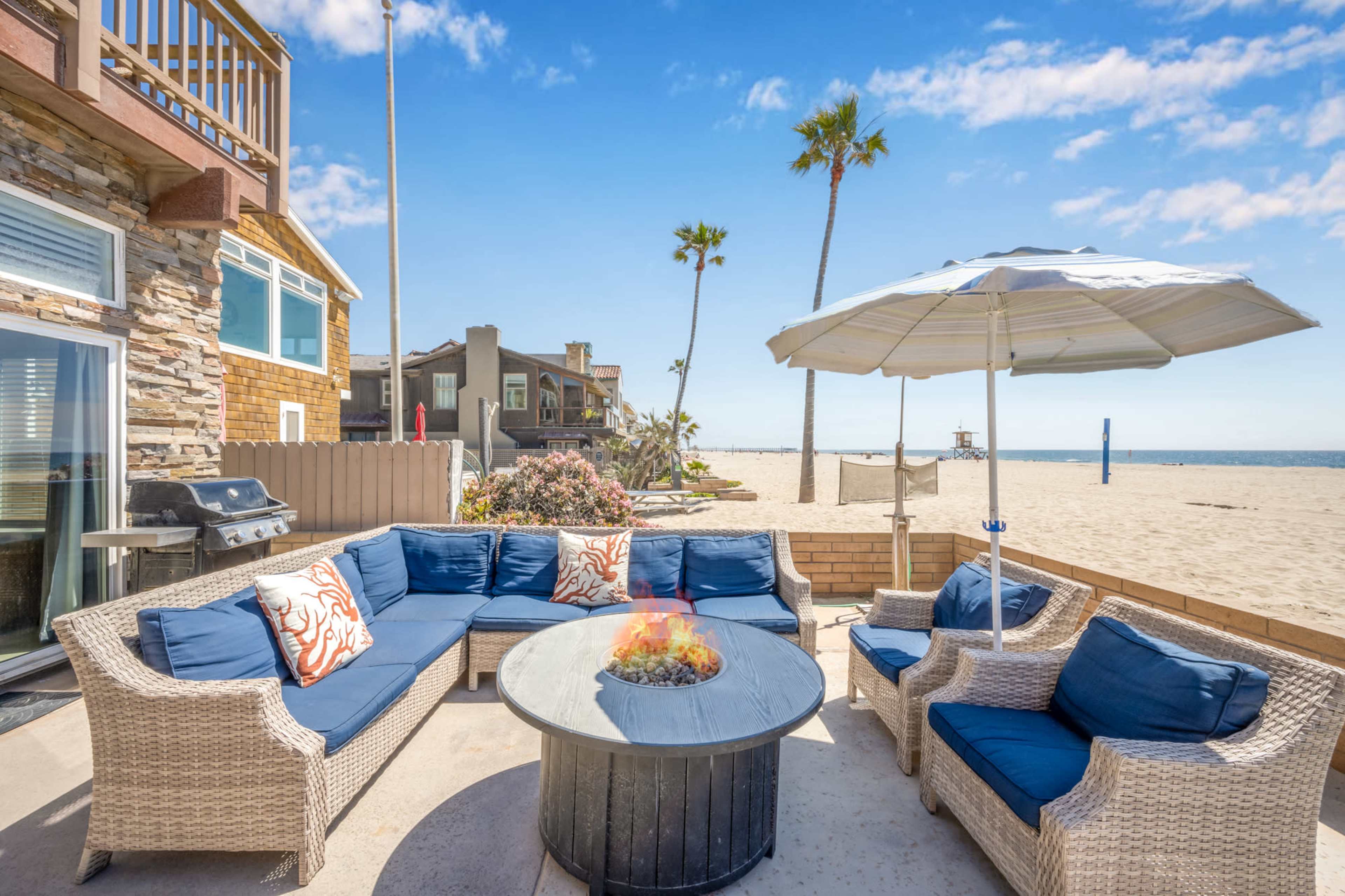 A patio area features a circular fire pit surrounded by a seating arrangement, overlooking a sandy beach and palm trees.