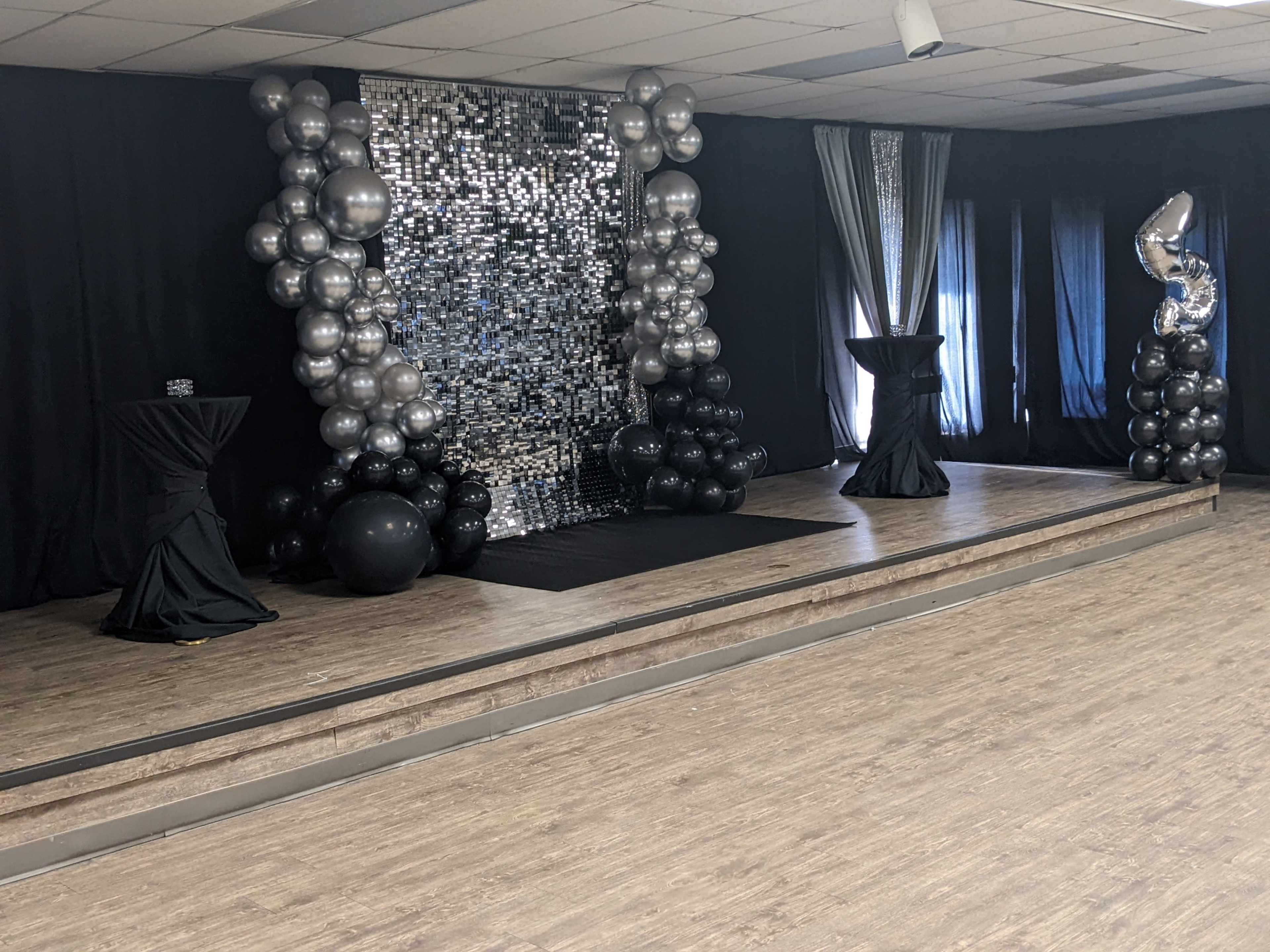 The image shows a decorated stage area featuring a shiny mirrored backdrop, balloon clusters in black and silver, and two draped tables.