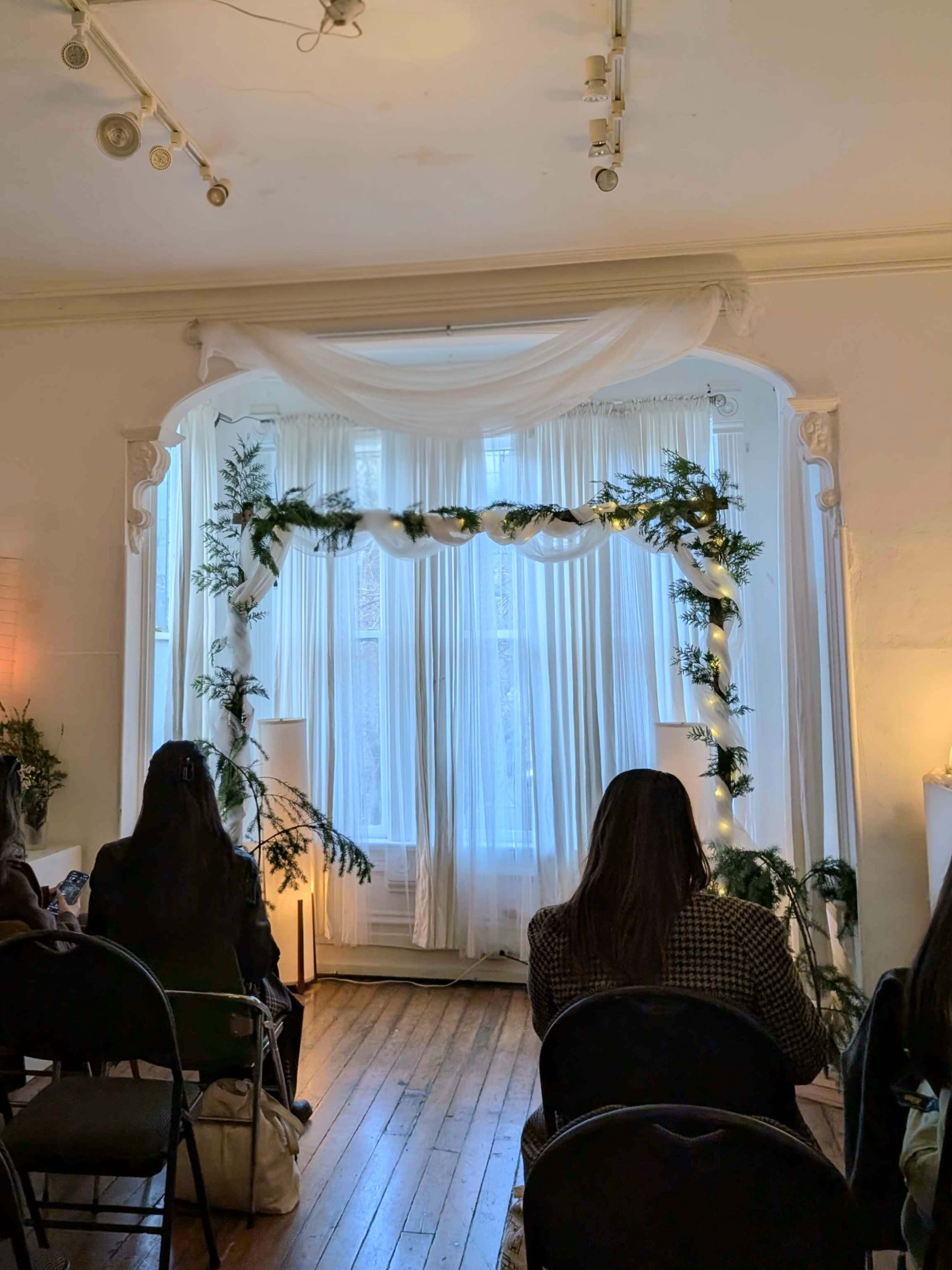 The image shows a room with a decorated arch made of greenery and fabric, set against a backdrop of light, flowing curtains.
