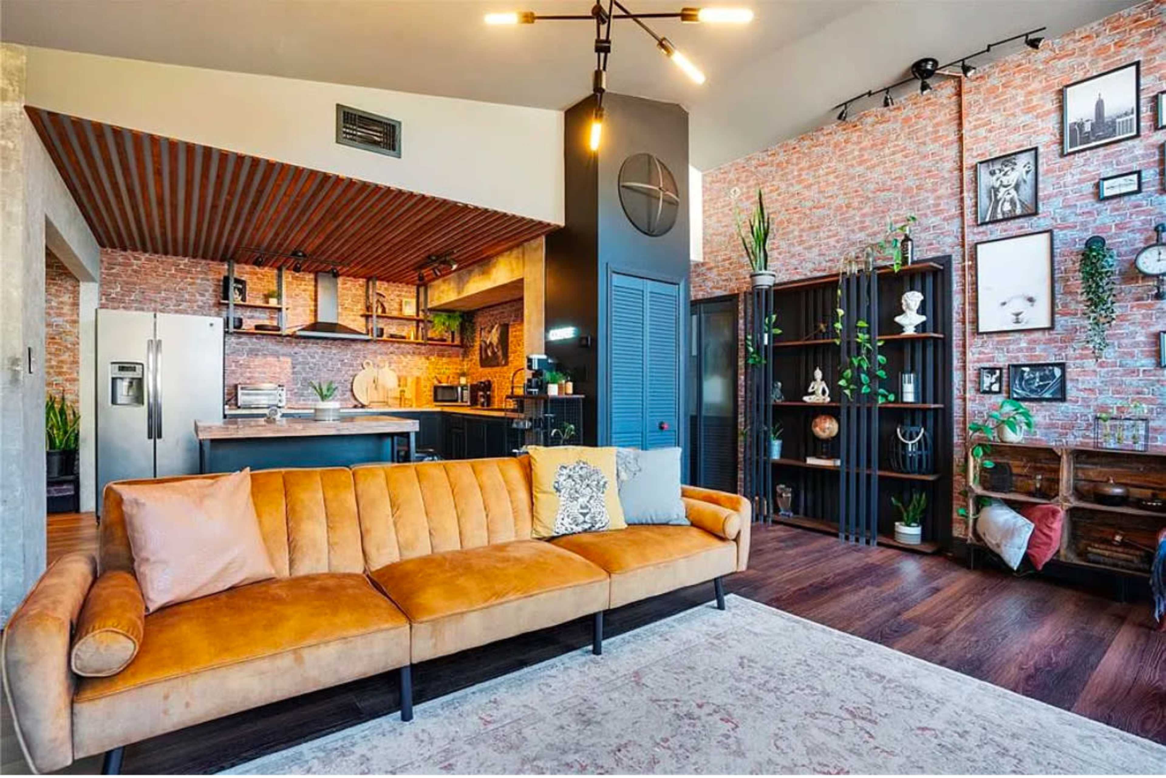 The image shows a modern living room featuring a tan sofa, a rustic kitchen area with exposed brick walls, and various decorative elements like plants and framed art.