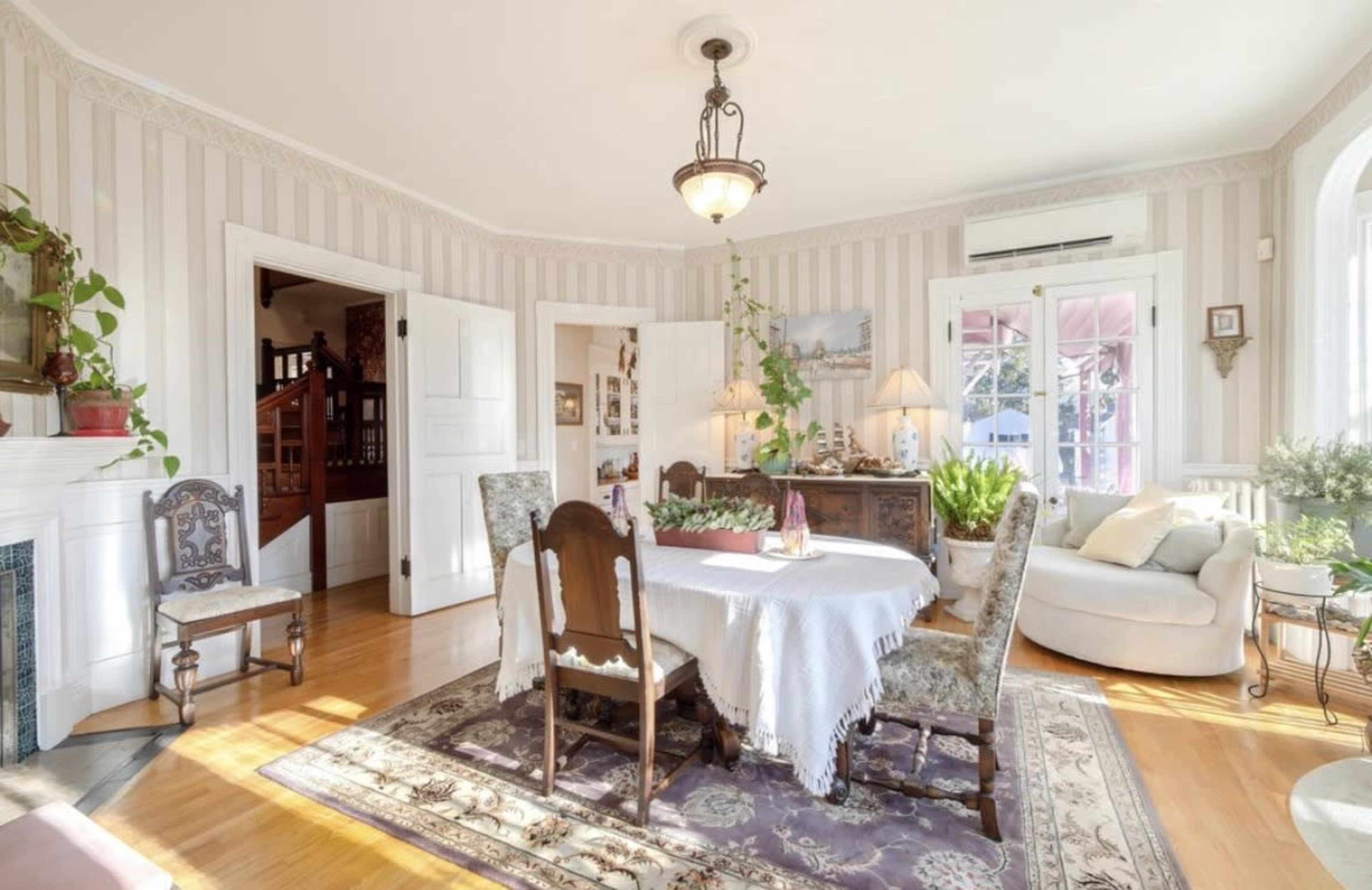 The image shows a bright dining area with a round table surrounded by chairs, accented by light-colored walls and natural light streaming in from large windows.