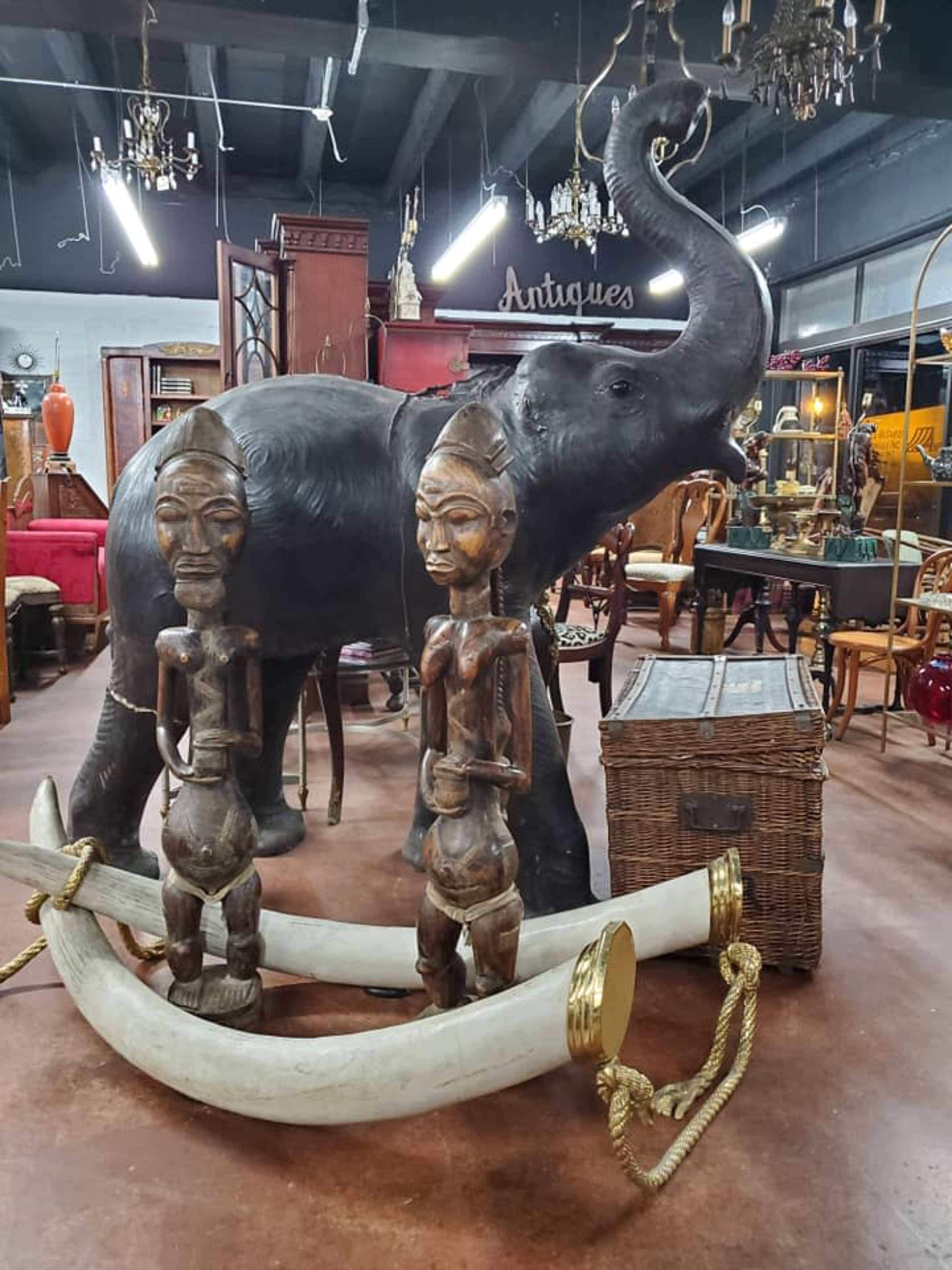 The scene features an antique shop displaying a large elephant sculpture alongside two wooden figures and a decorative piece resembling curved tusks.