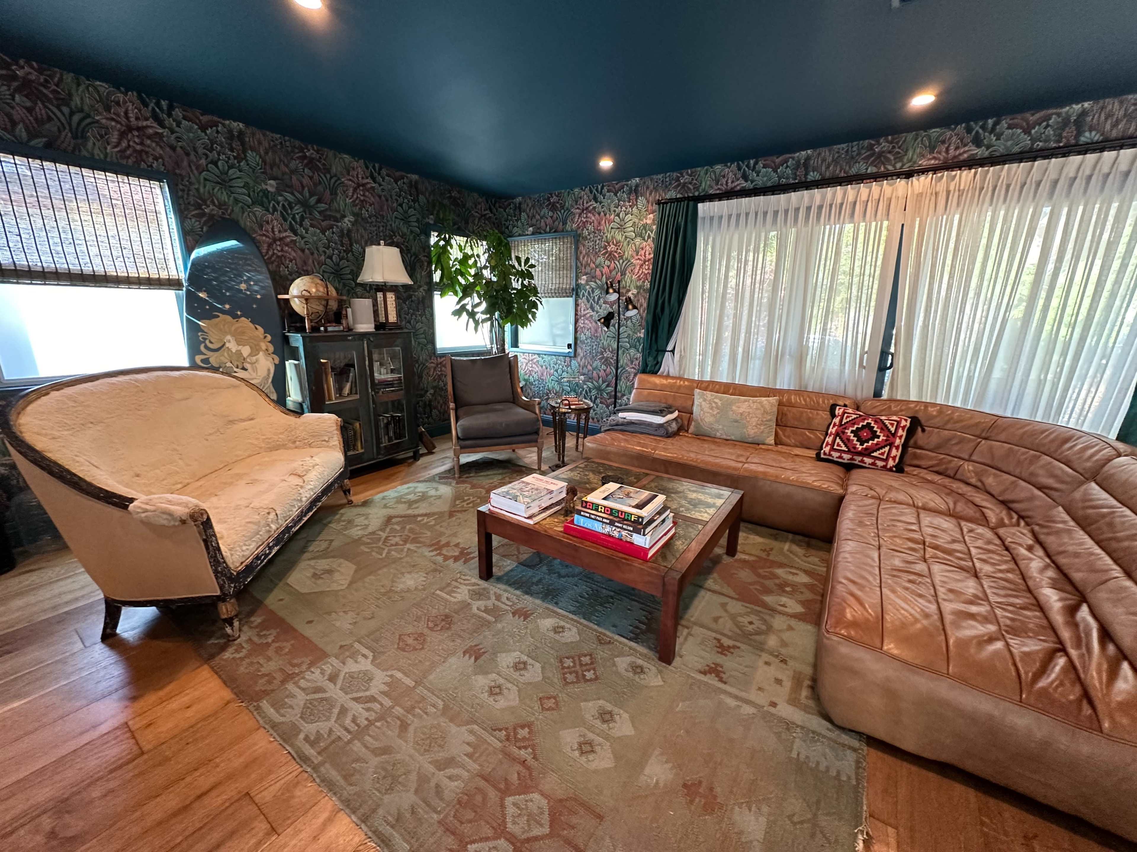 A cozy living room features a sectional leather sofa, a circular chair, a wooden coffee table stacked with books, and wallpaper adorned with floral designs.