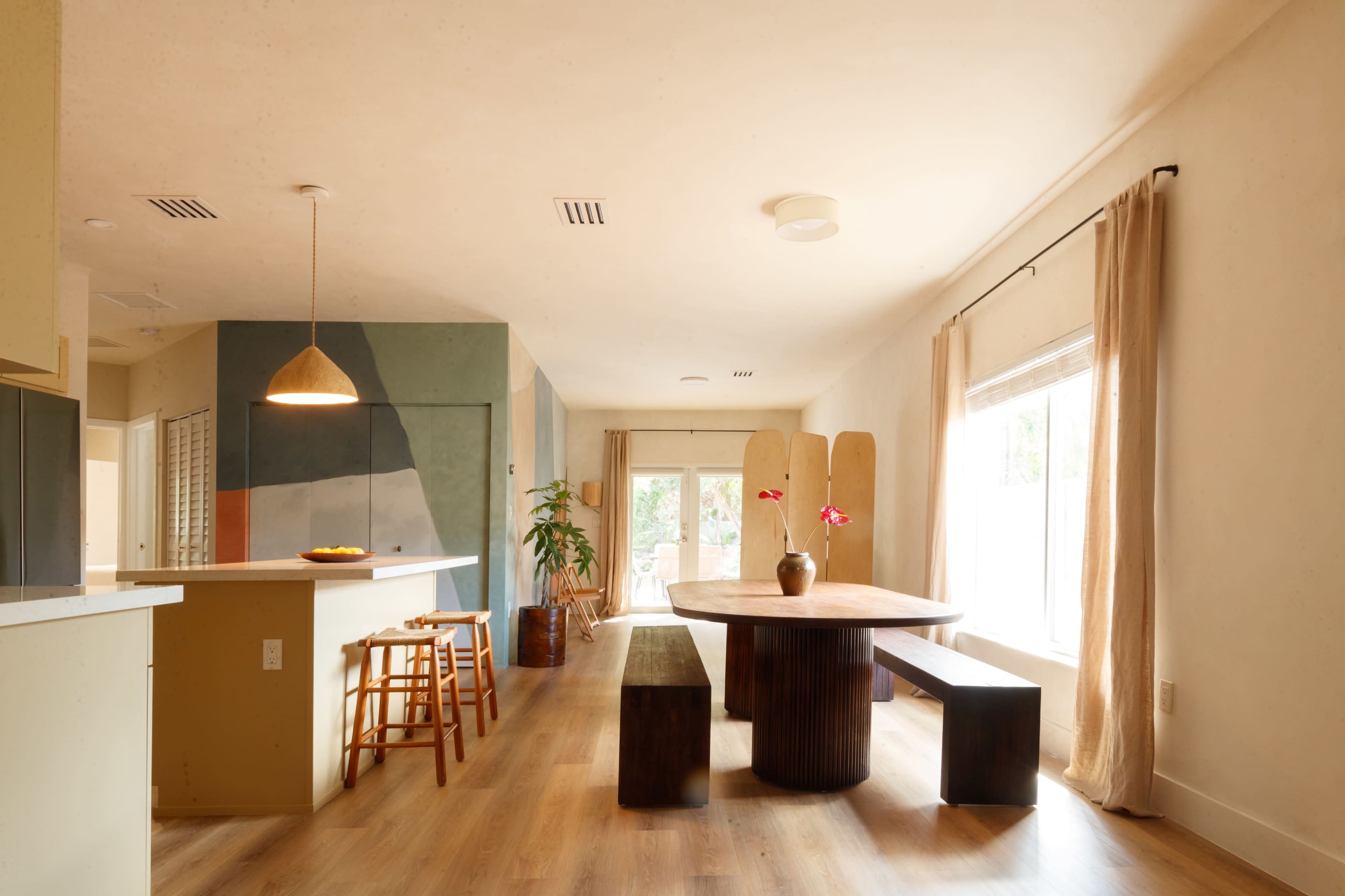 The image depicts a modern interior dining area featuring a round wooden table surrounded by two rectangular benches, with large windows allowing natural light to enter the space.