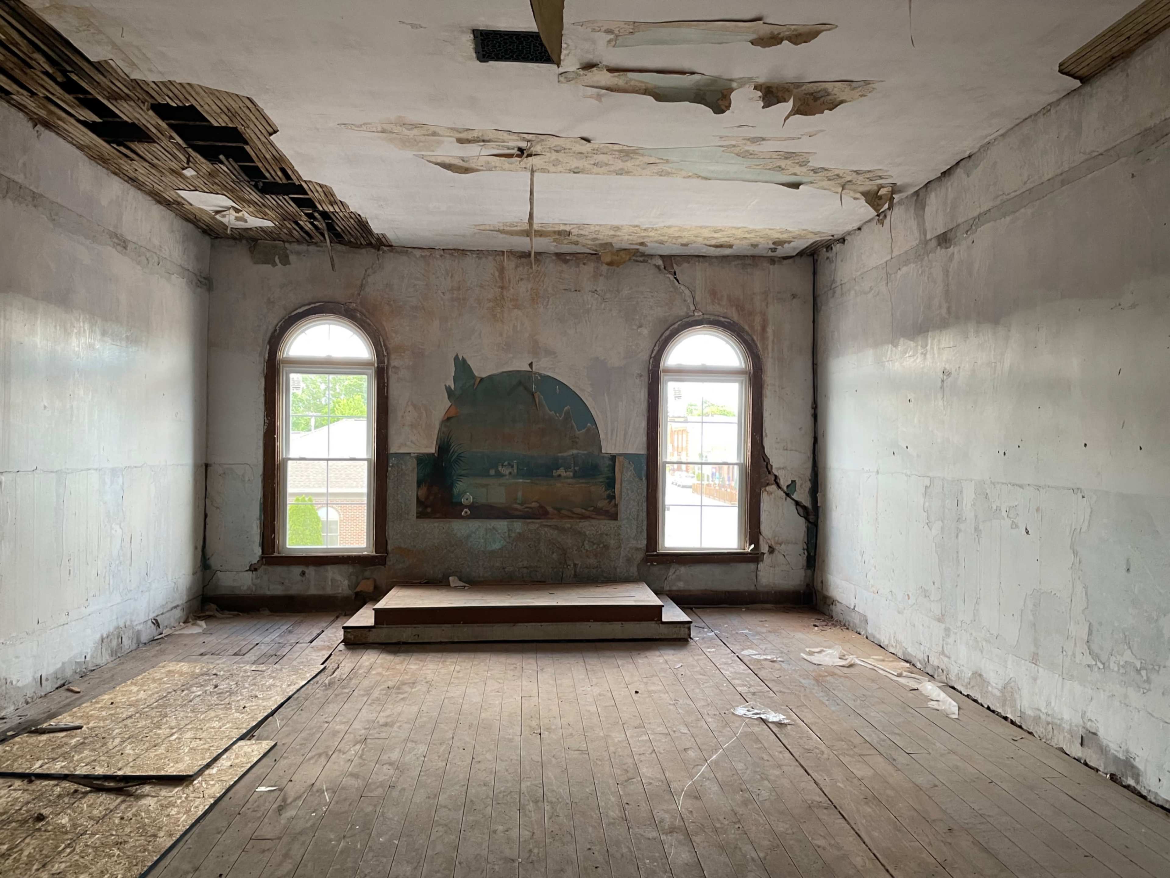 The image shows an empty, dilapidated room with peeling walls, two arched windows, and a small raised platform in the center.