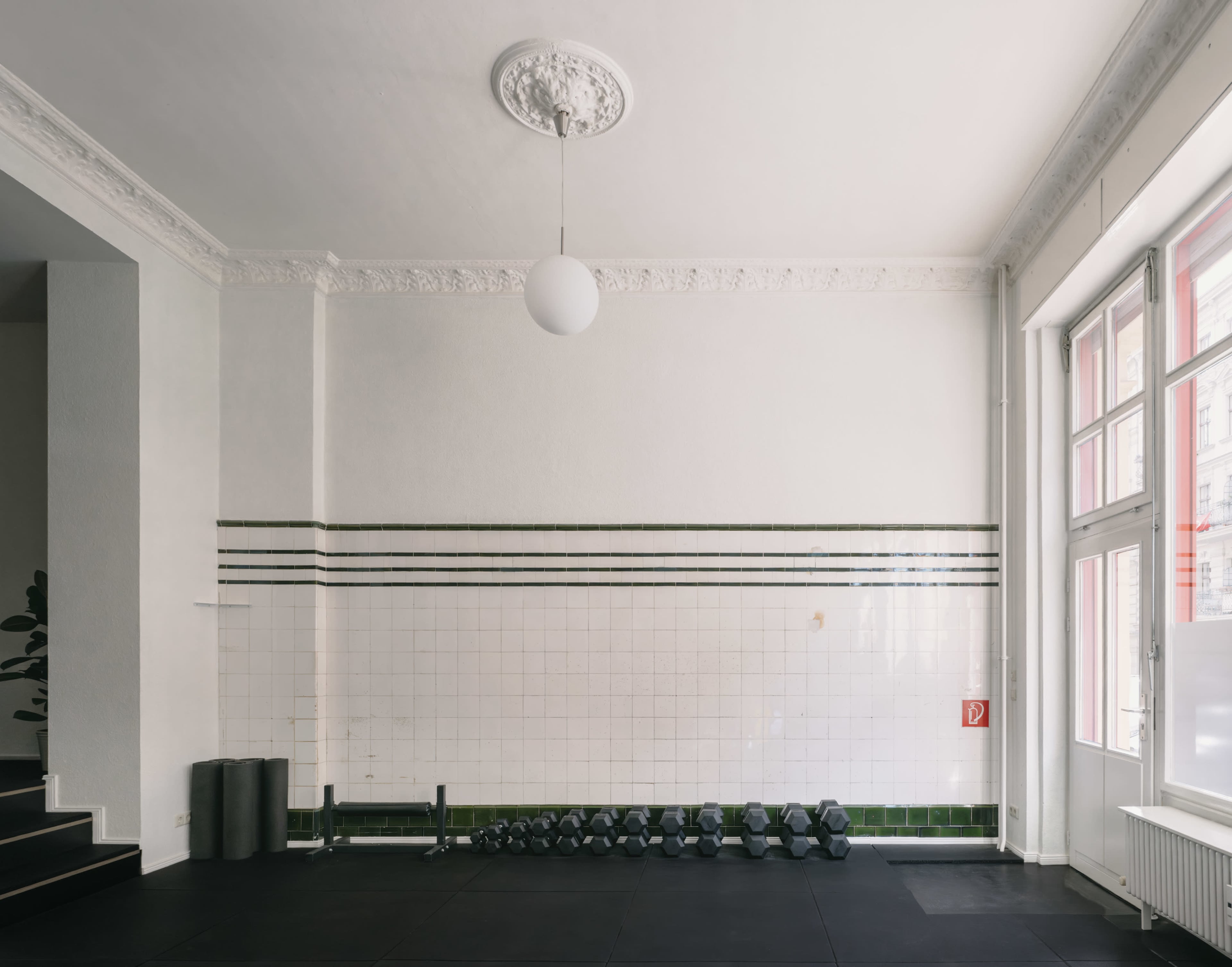 The image shows a spacious room with a tiled wall, featuring a row of dumbbells on the floor and large windows allowing natural light.