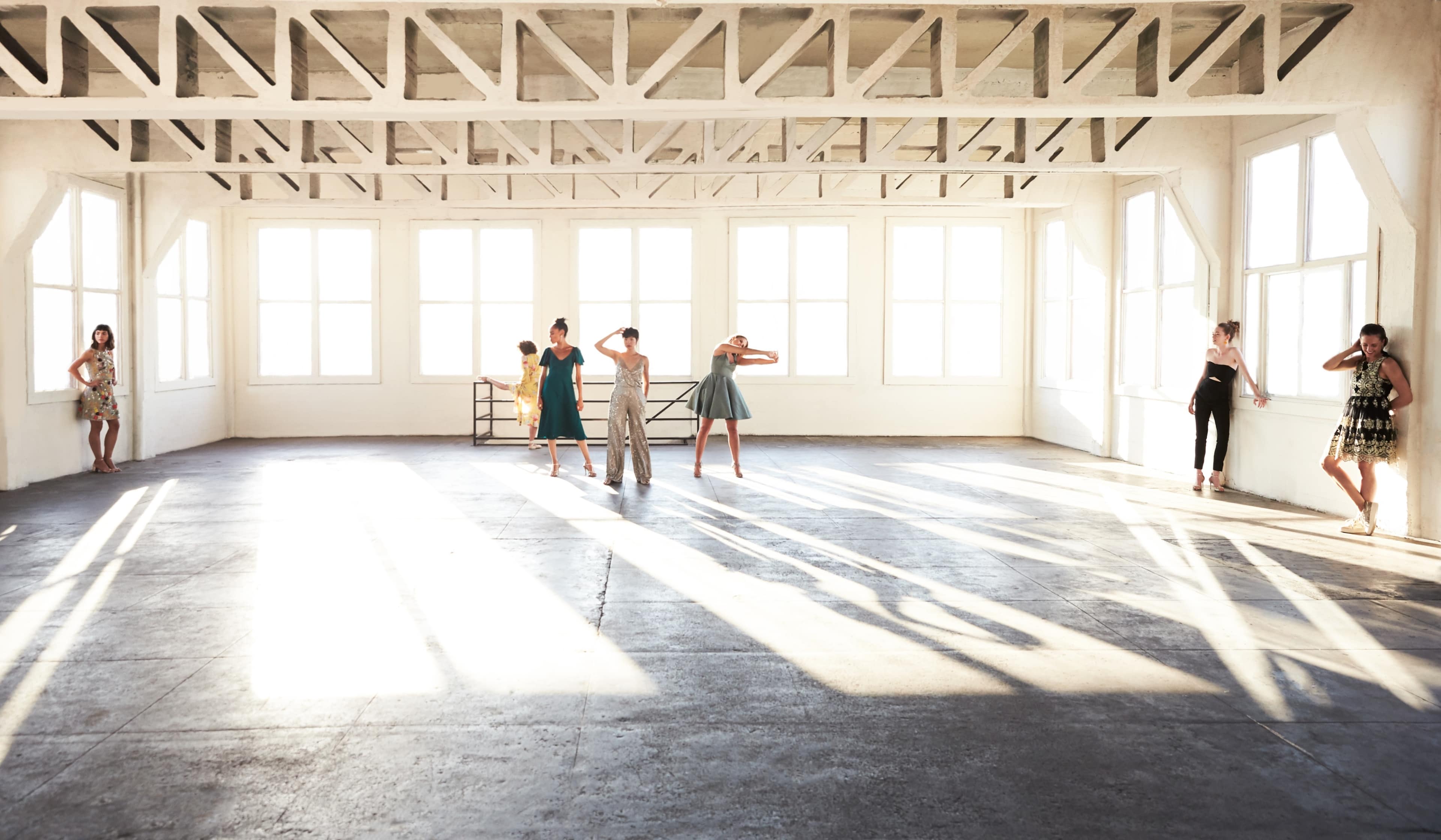 A group of women stands in various poses in a sunlit, spacious room with large windows.