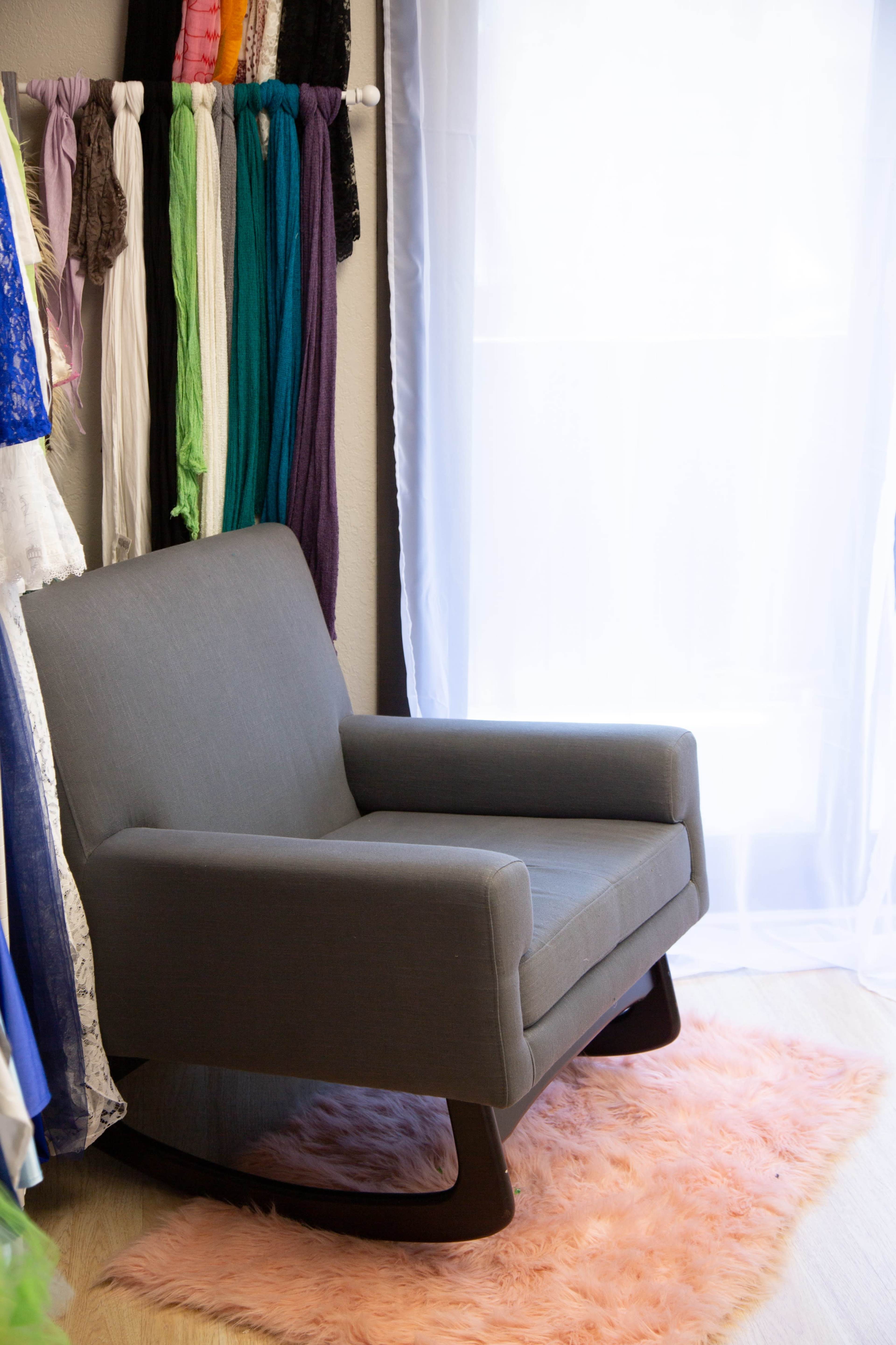 A gray chair is positioned next to a window with sheer curtains, surrounded by colorful fabrics hanging on a rod and resting on a pink shag rug.