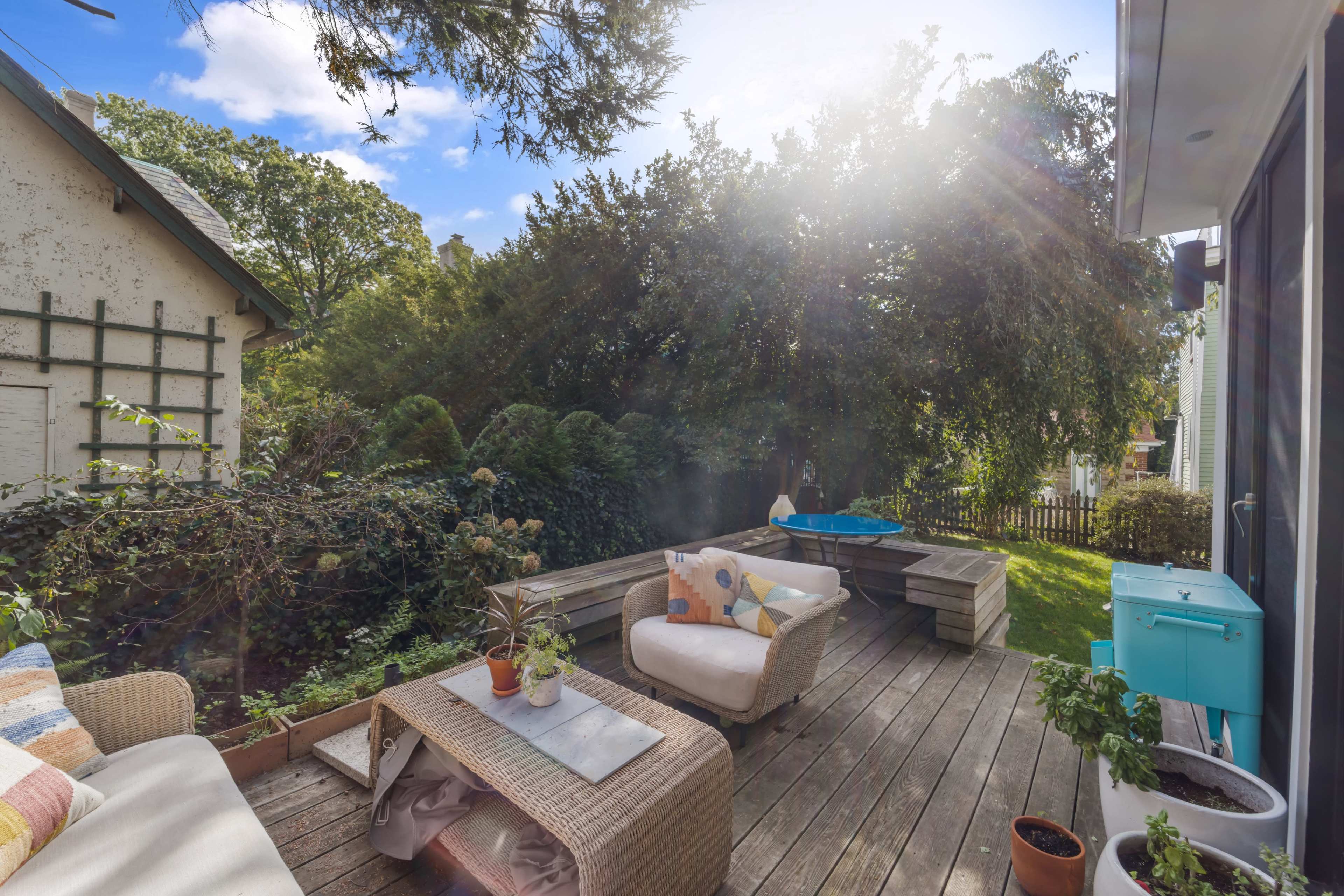 A wooden patio features a seating area with a couch and coffee table, surrounded by greenery and a bright sky in the background.