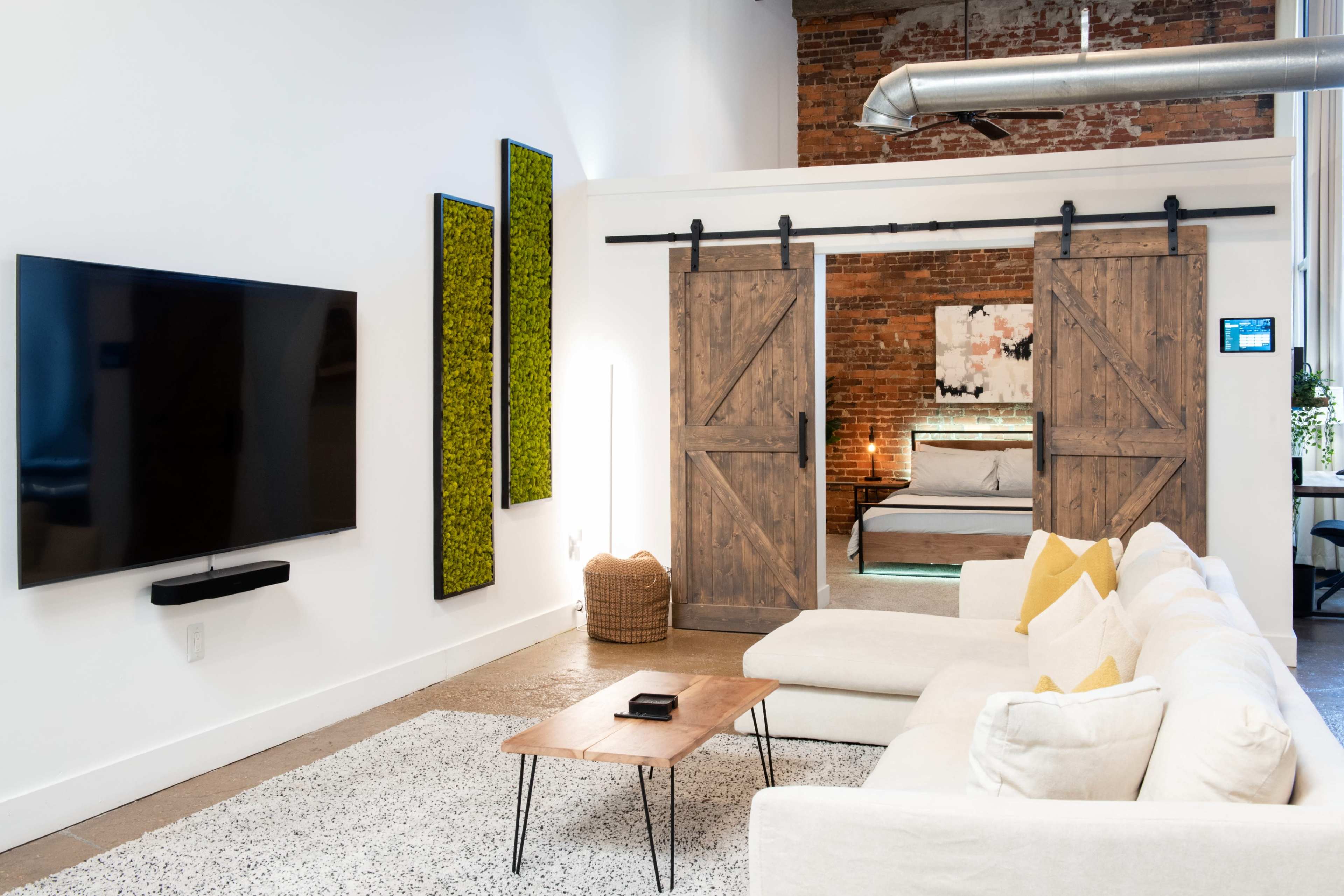 A modern living room features a large TV mounted on the wall, a white sectional sofa, a wooden coffee table, and sliding barn doors leading to a bedroom area with exposed brick walls.