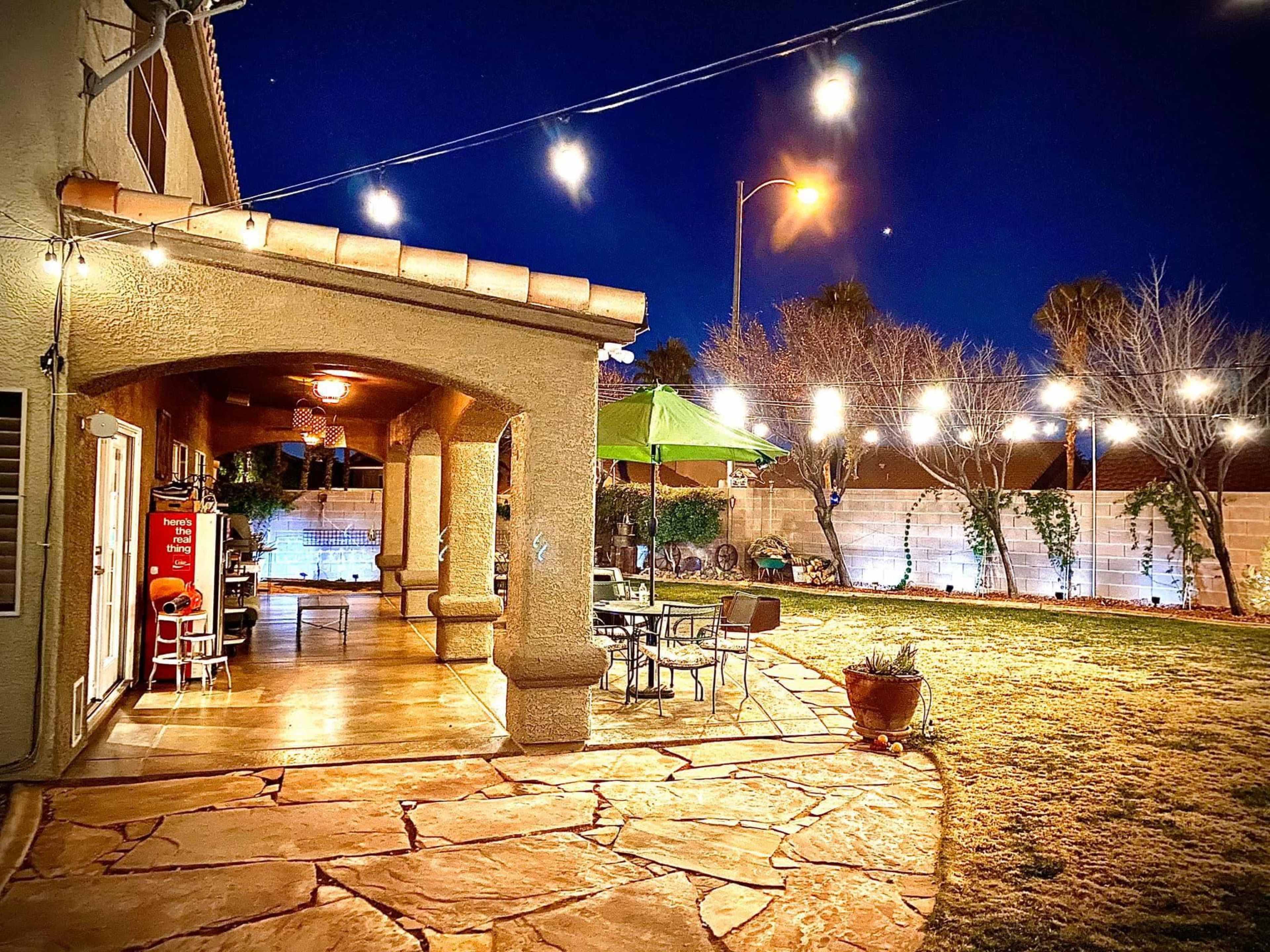 A well-lit backyard patio with string lights illuminating a stone pathway, surrounded by a green lawn and a house.