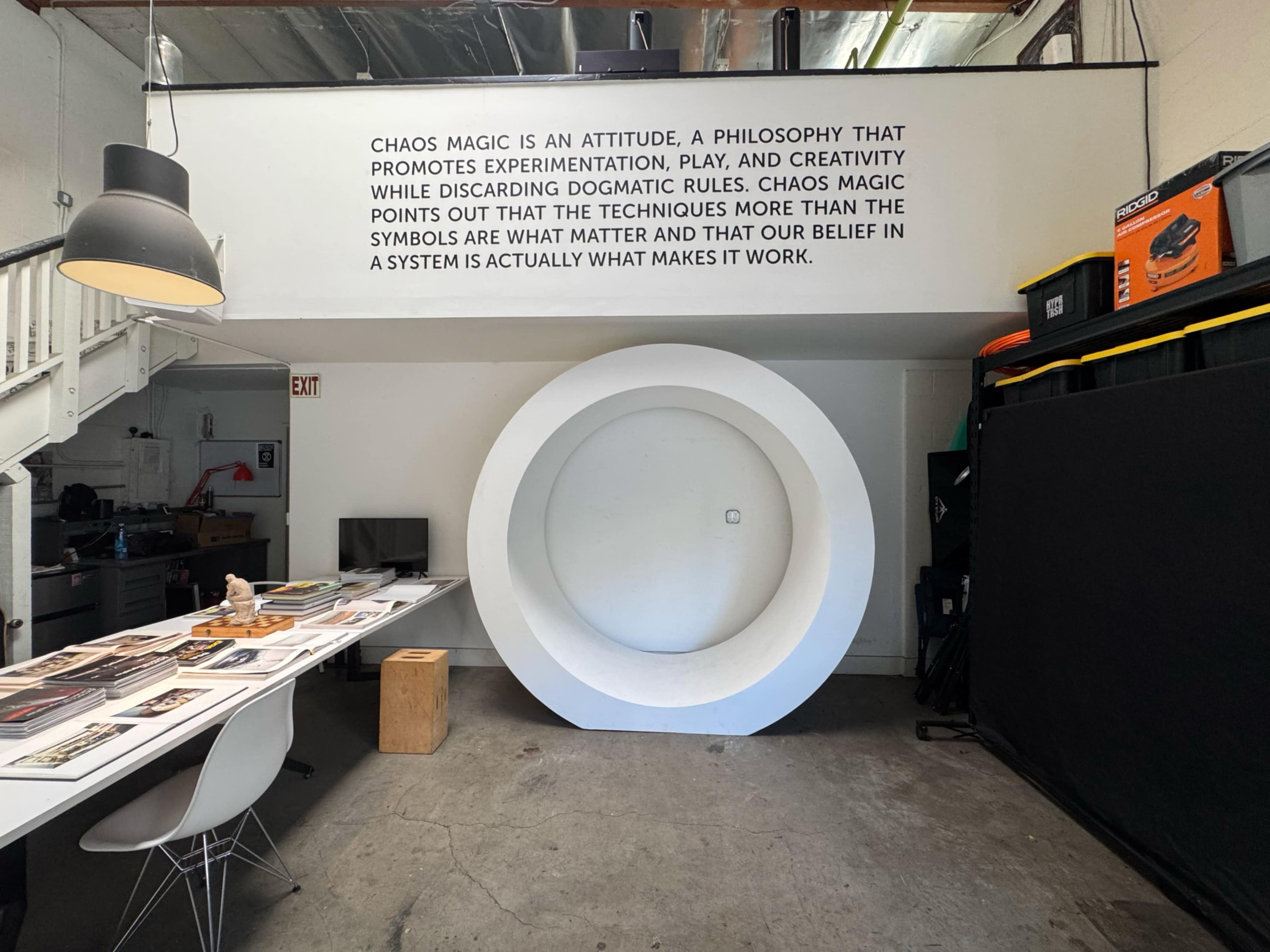 The image shows an interior space featuring a circular white installation on the wall, with a table and chairs arranged nearby, and text about chaos magic displayed above.
