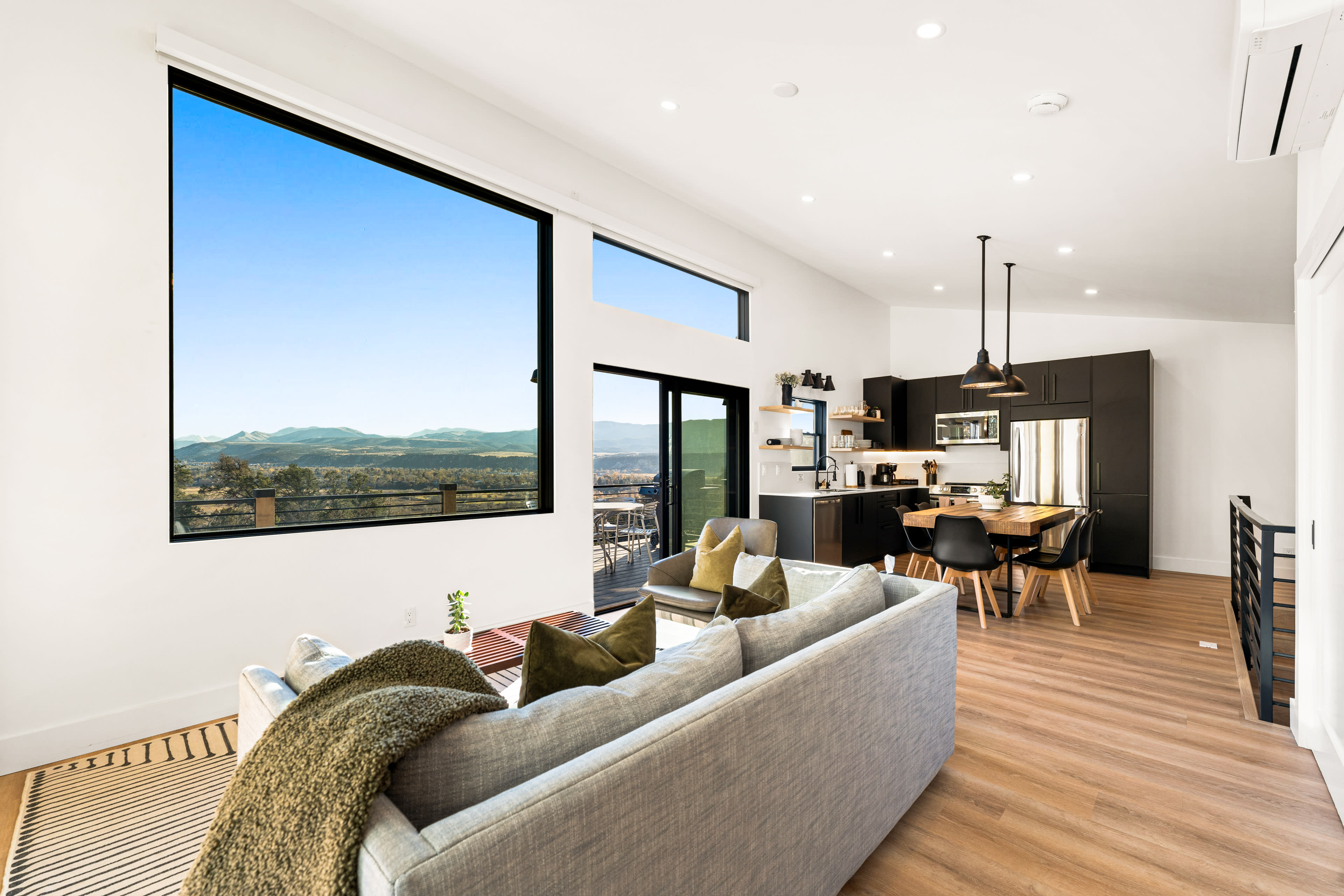 The image shows a modern living area with a large window overlooking mountains, featuring a gray sofa, wooden flooring, and a stylish kitchen in the background.