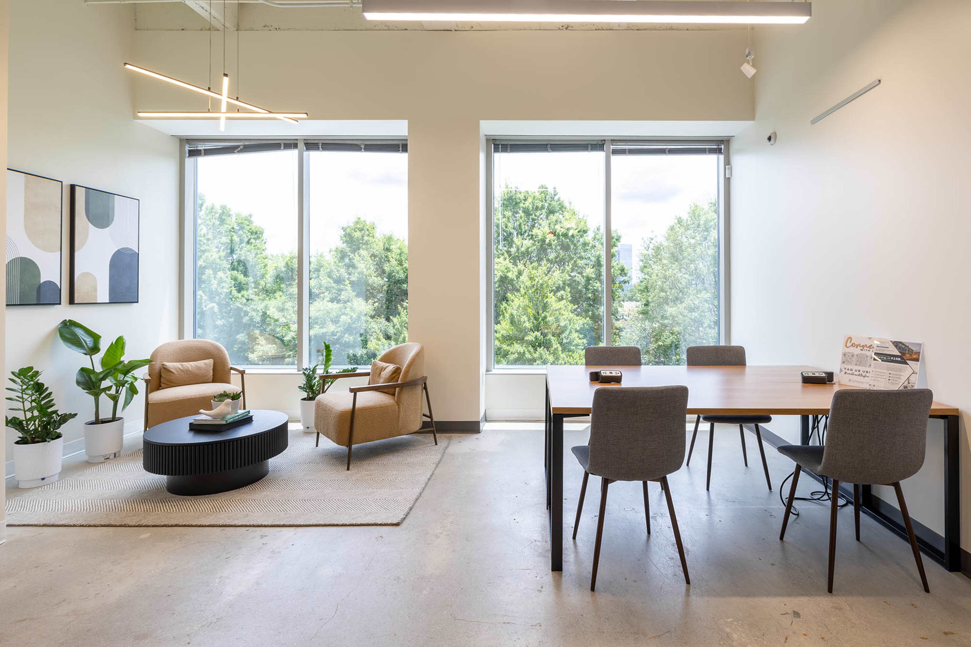 The image shows a modern office space featuring a seating area with two chairs and a coffee table, and a dining table with four chairs, all surrounded by large windows overlooking greenery.