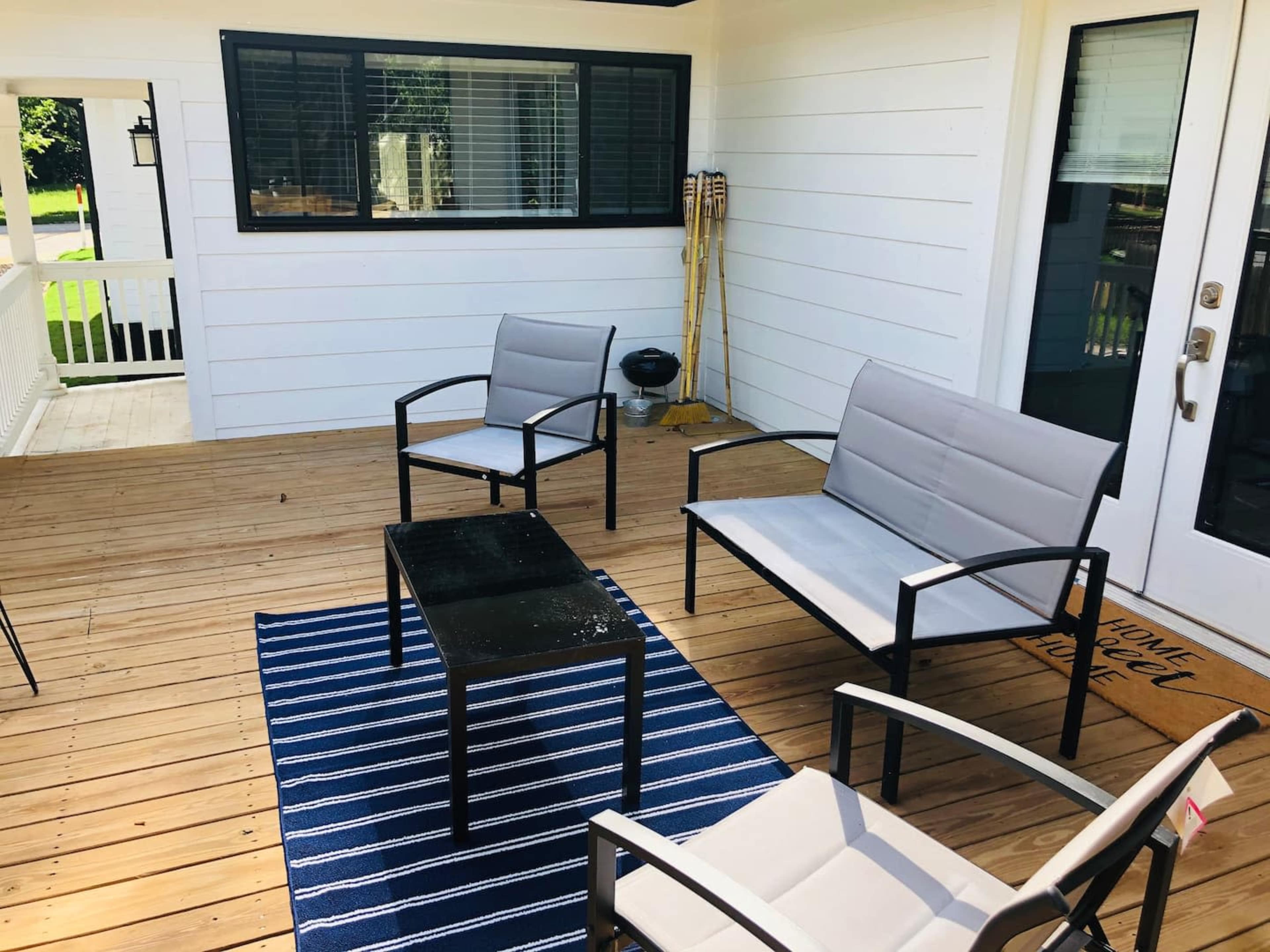 A porch with a wooden floor, two gray chairs, a black table, and a blue striped rug.