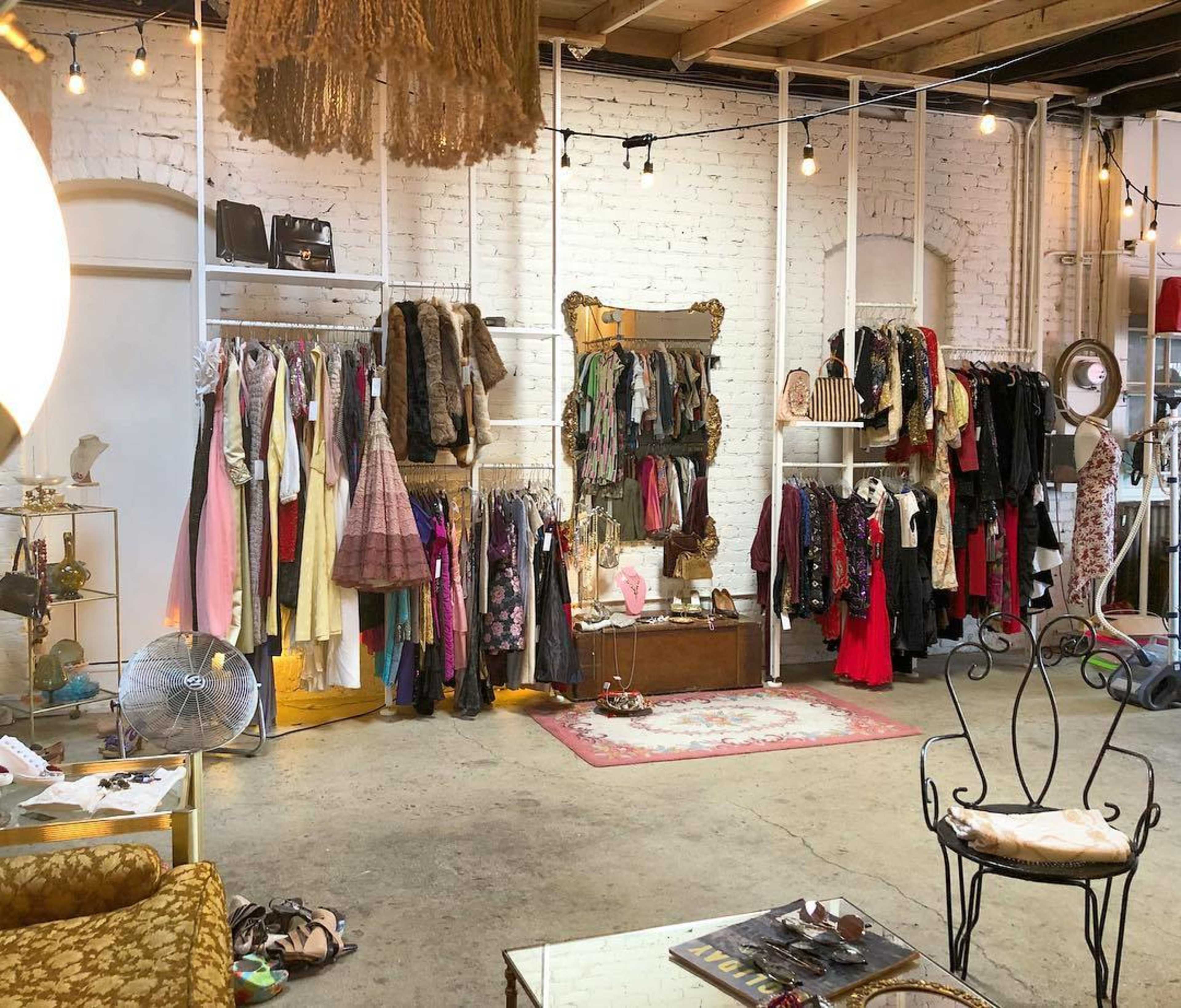 A vintage clothing store interior with racks of assorted garments and accessories arranged on white brick walls, along with seating and decorative items.